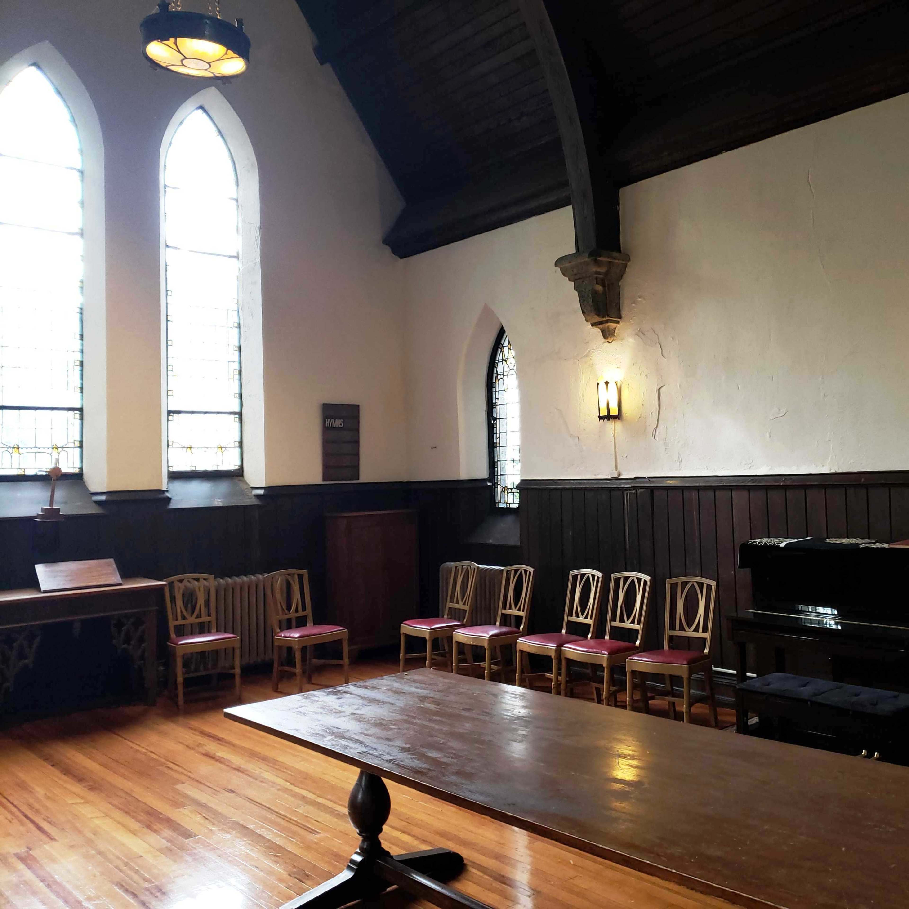 The interior of a church features a wooden table in the foreground, seven chairs arranged along one wall, and a black piano against another wall, illuminated by light from stained glass windows.