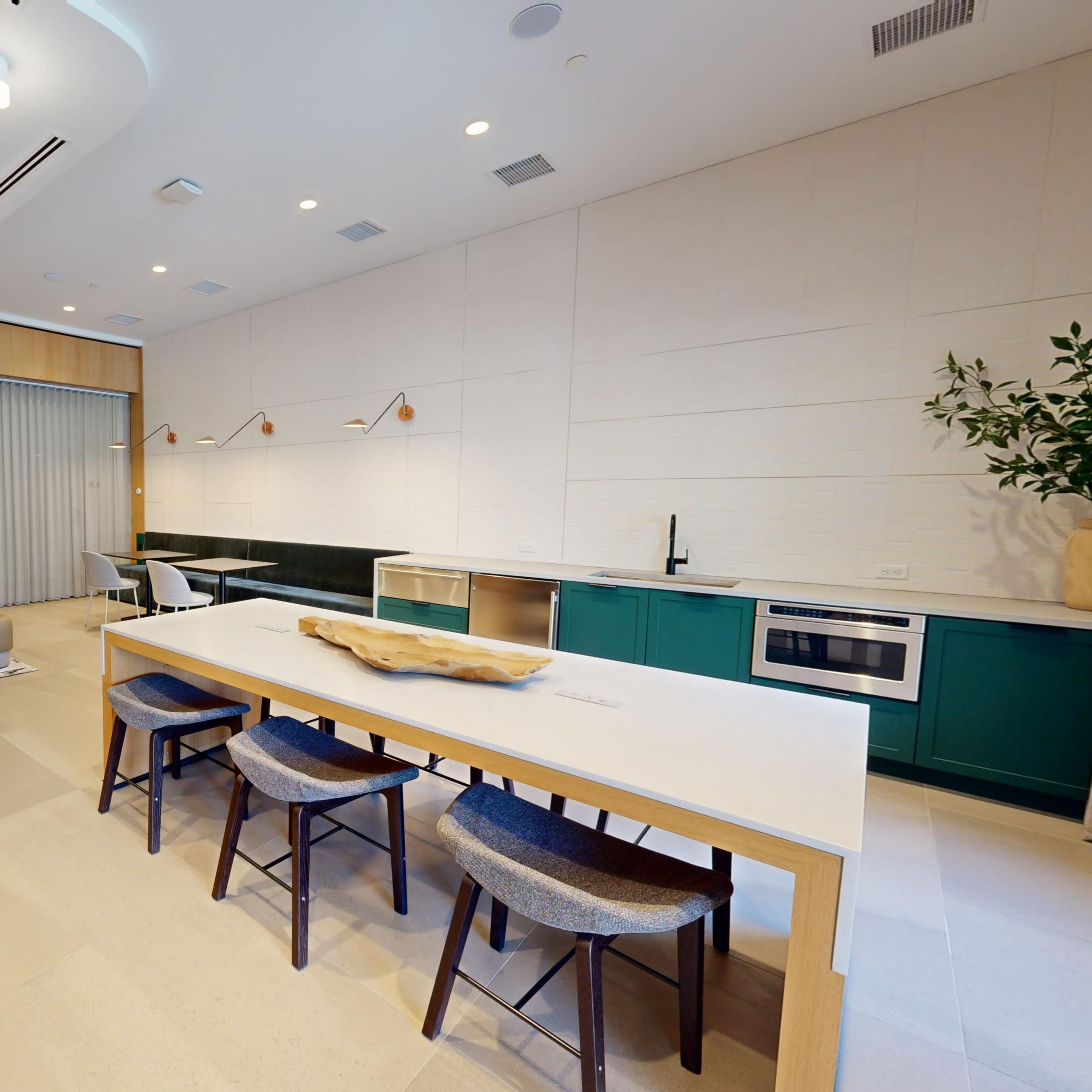 A modern living space featuring a dining area with a large table, chairs, a sofa, a television mounted on a wall, and a minimalist kitchen in the background.
