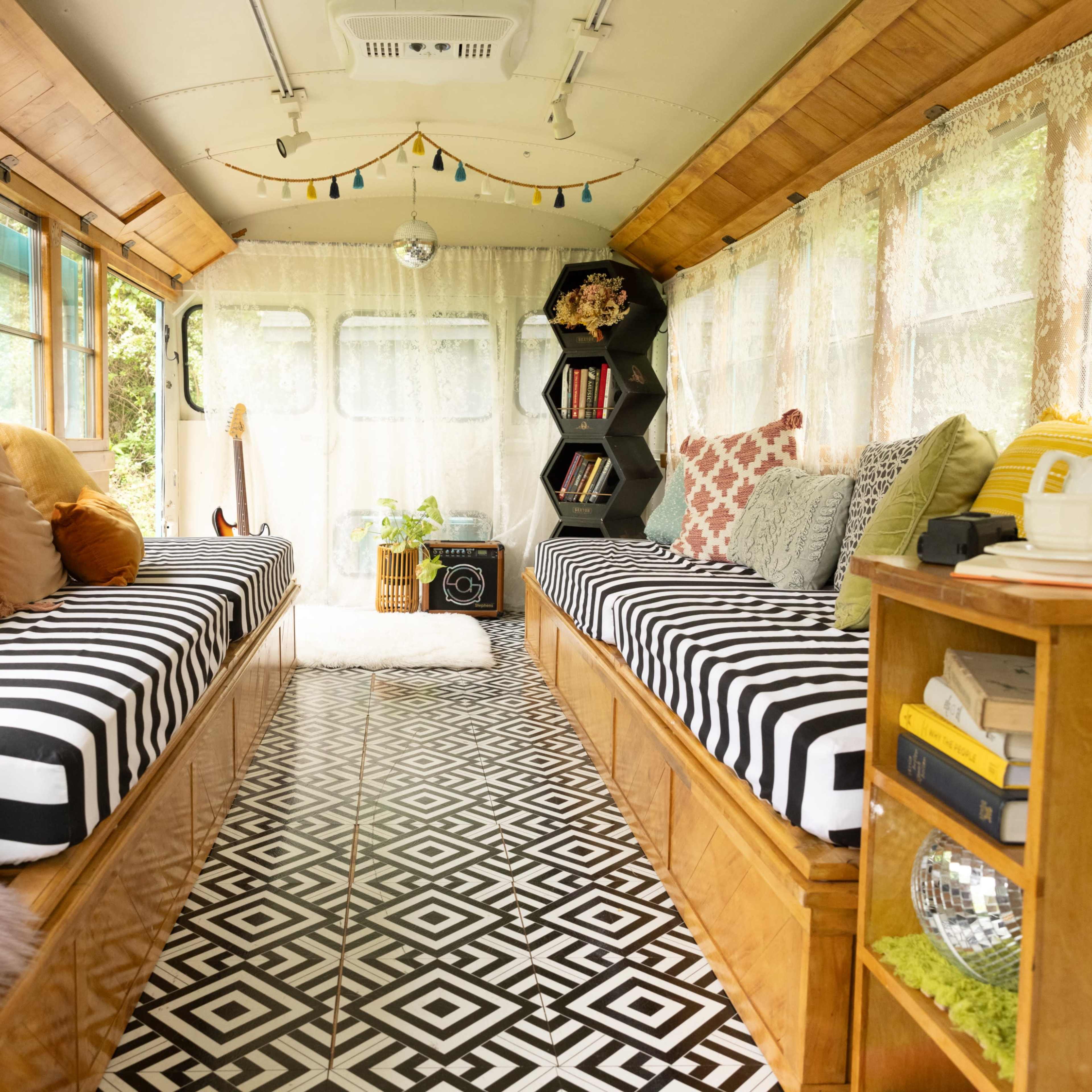 The interior of a renovated bus, featuring striped benches with colorful cushions, a patterned floor, and a bookshelf positioned beside a large window.