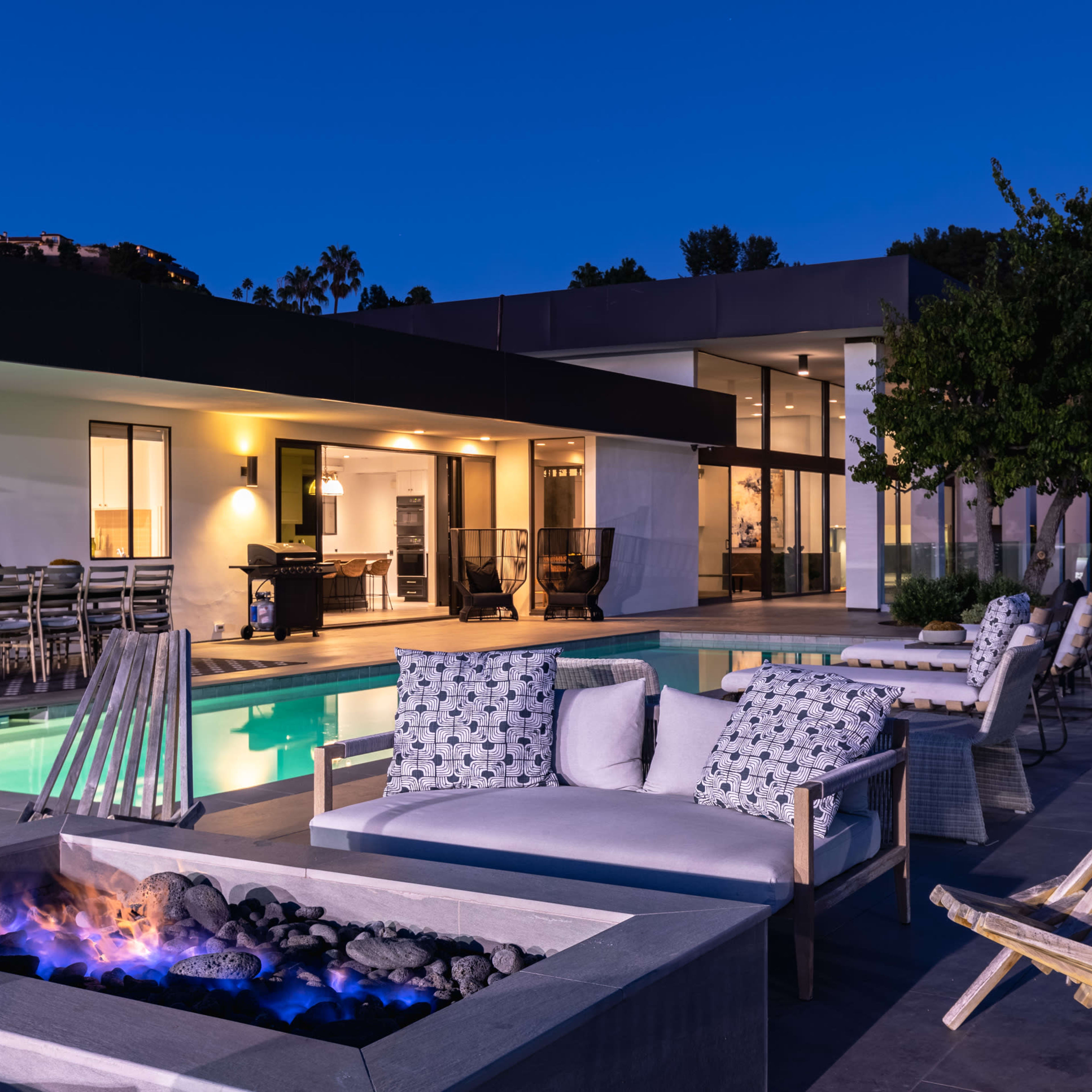 A modern backyard features a swimming pool, cozy seating around a fire pit, and a stylish house in the background, illuminated at night.