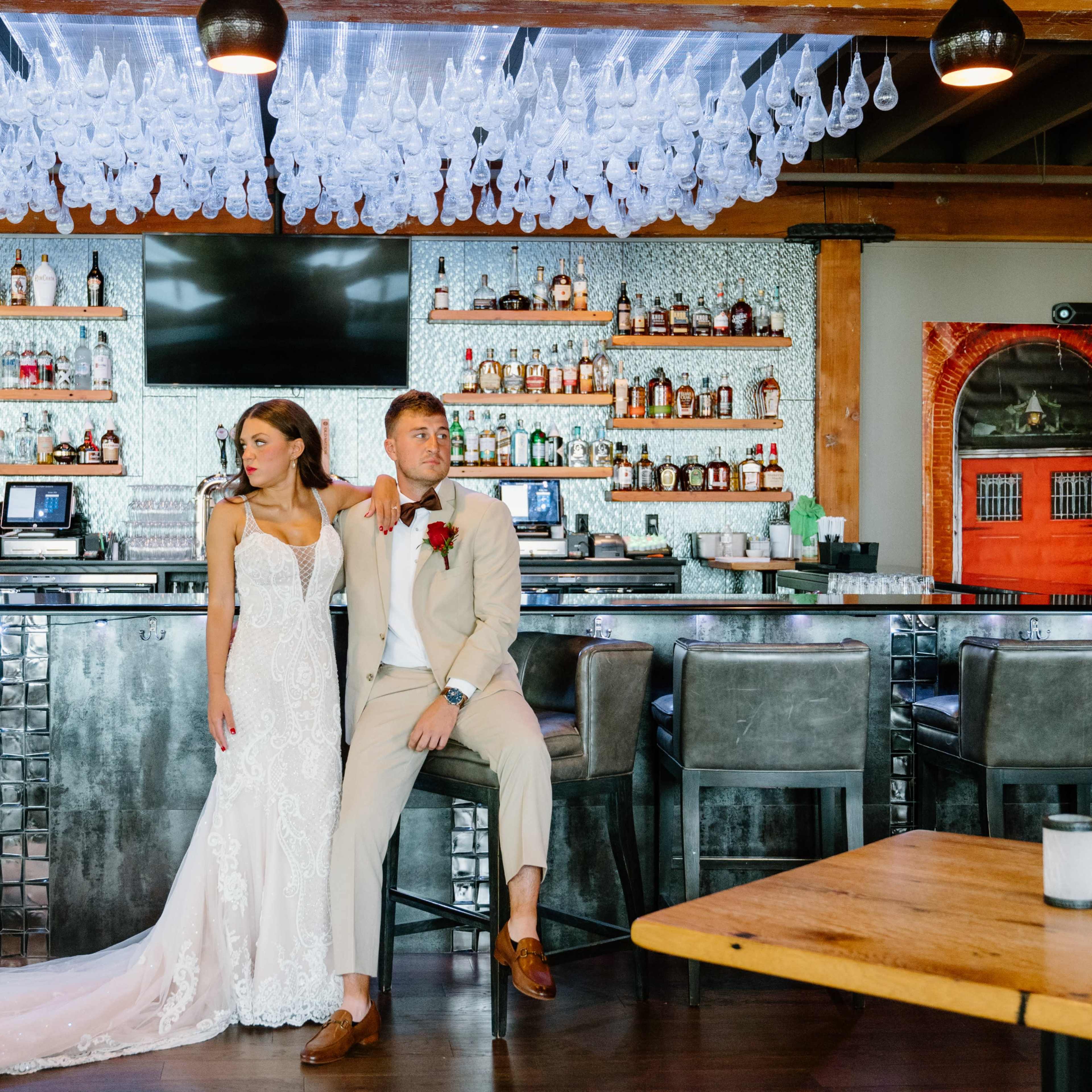 A bride in a lace wedding dress stands beside a groom in a beige suit at a bar with an array of bottles displayed on shelves behind them.