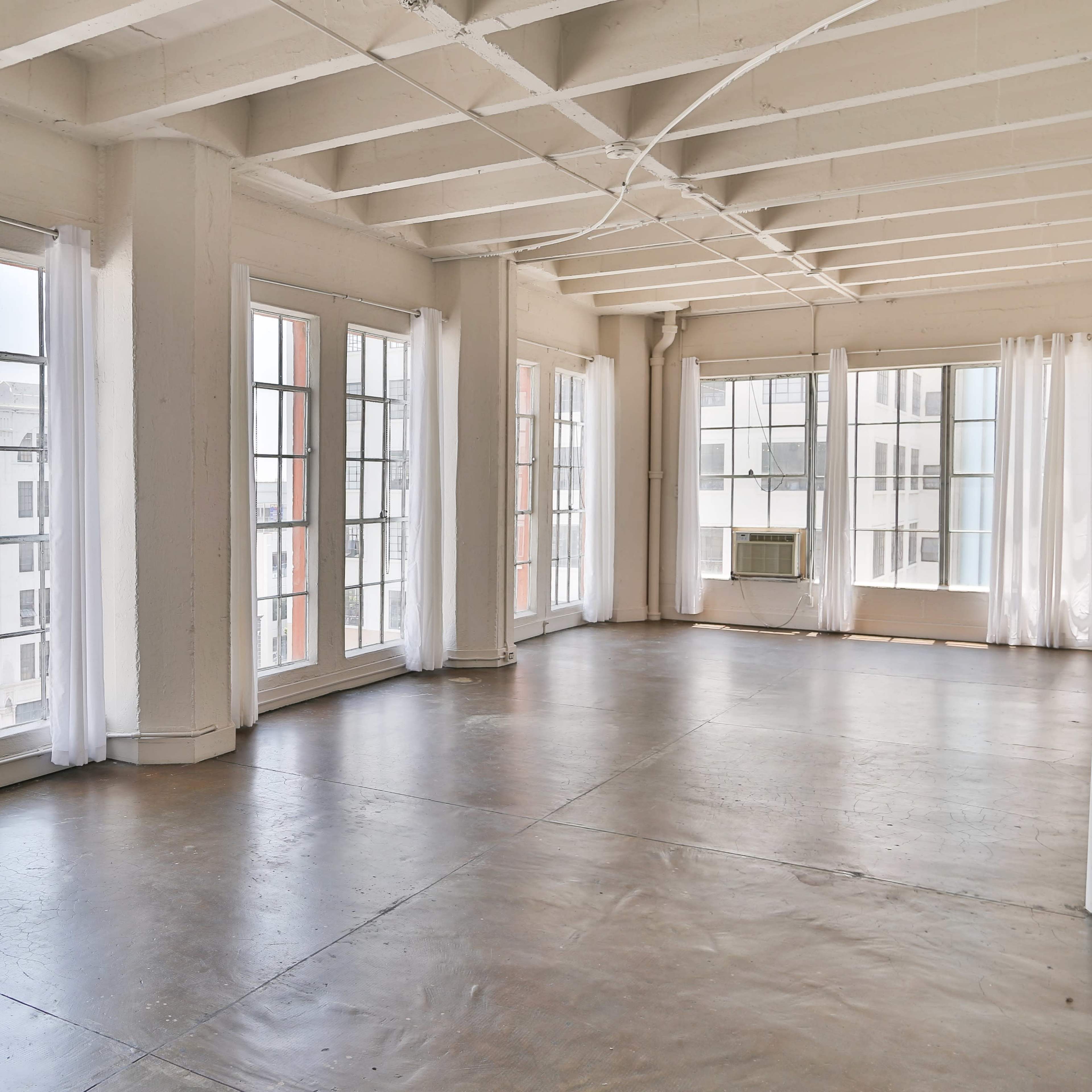 A spacious, empty room with large windows and polished concrete floors, featuring white walls and light curtains.