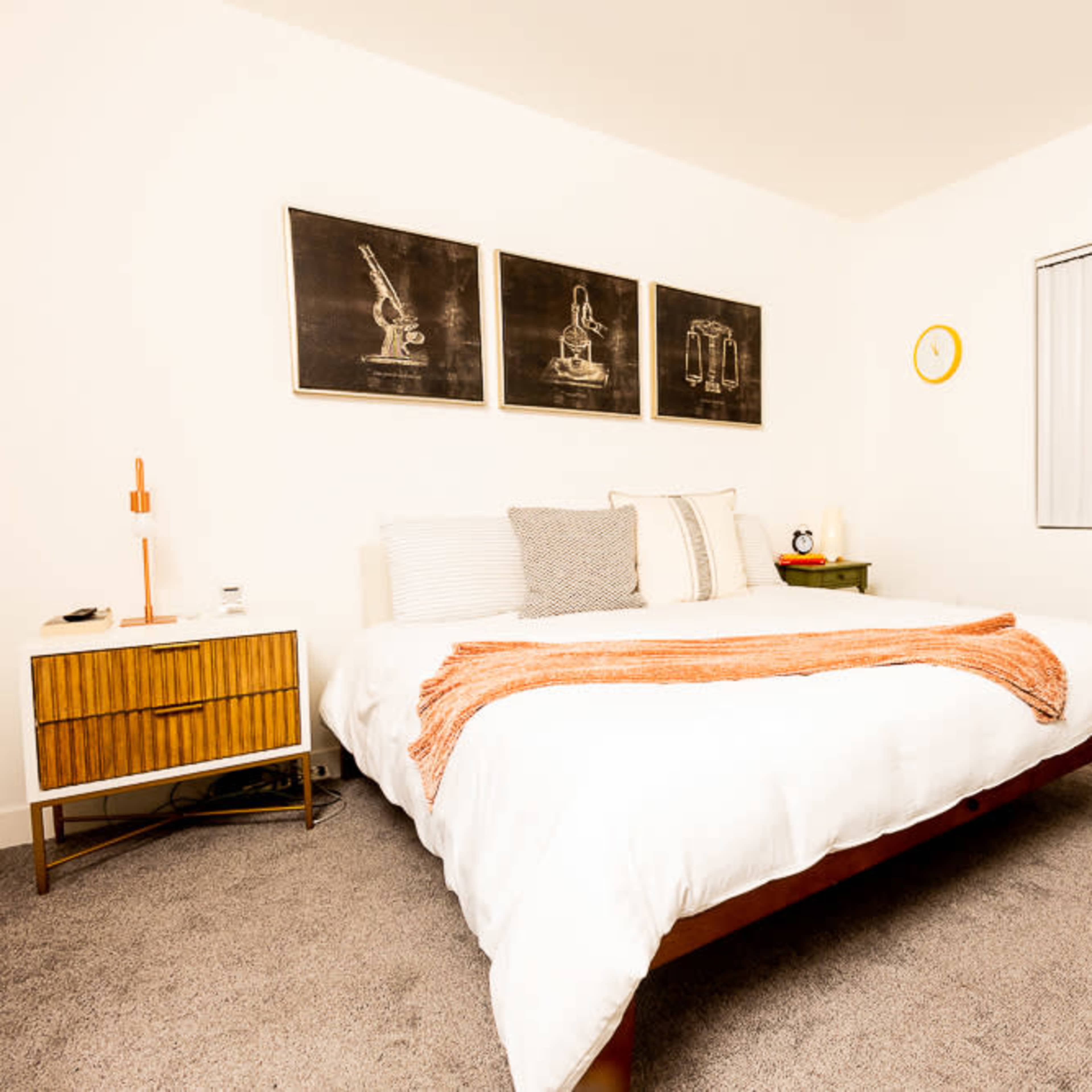 A well-organized bedroom features a bed with a white comforter, two side tables, and framed artwork on the walls.