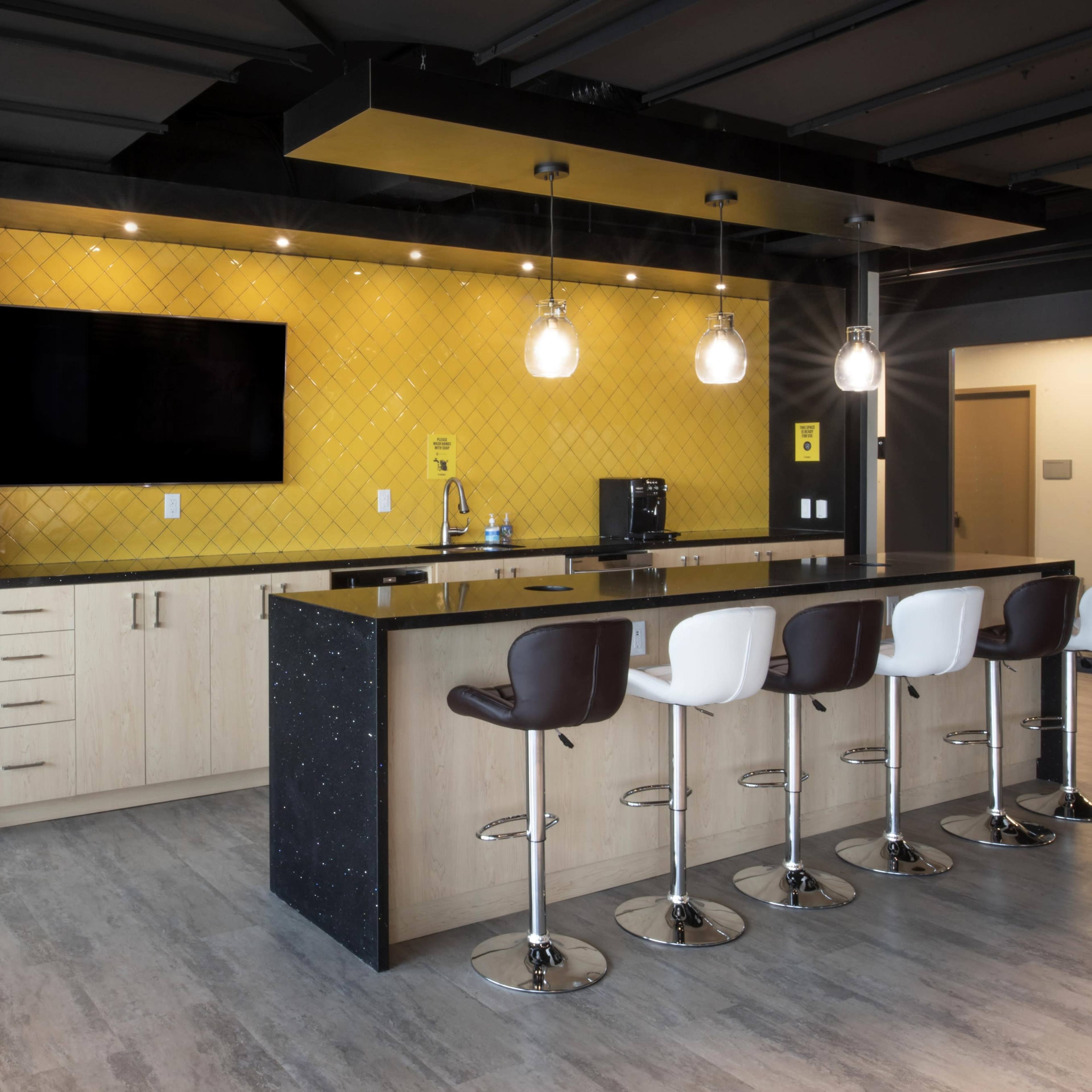A modern kitchen area in an office with a long bar counter, four high stools, a television mounted on the wall, and yellow tiles as a backdrop.