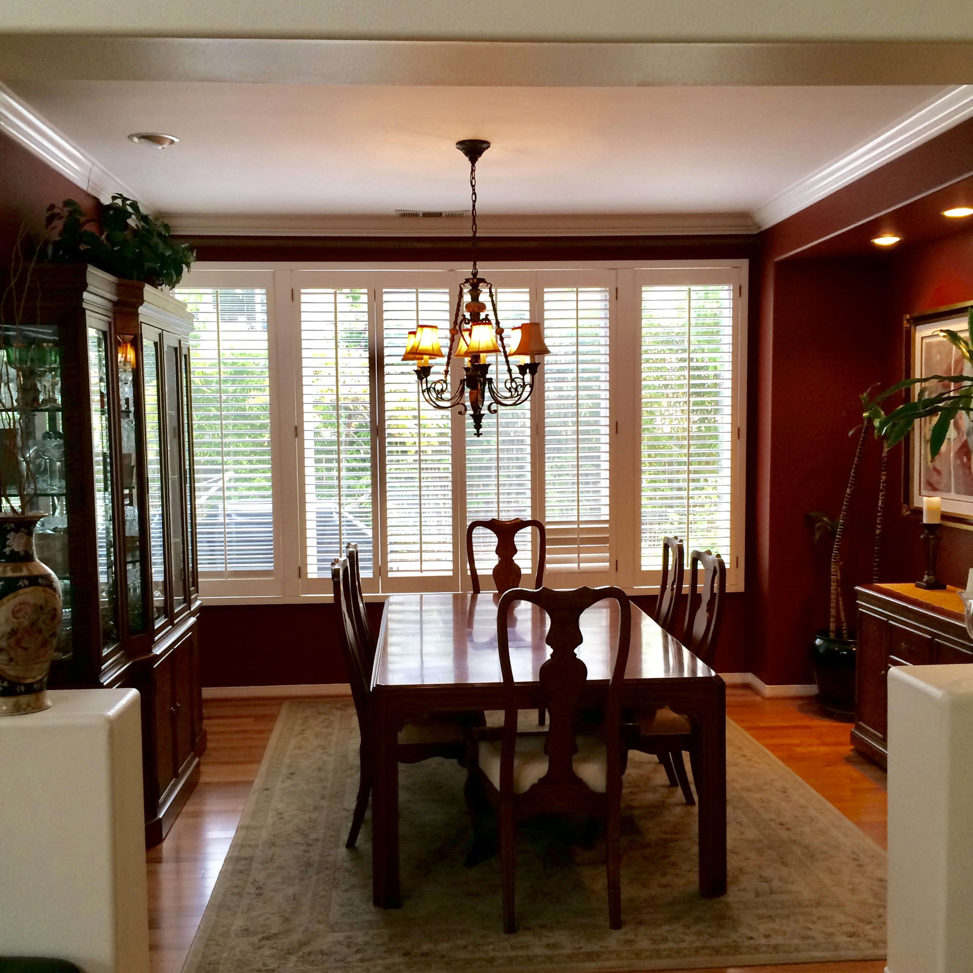 A dining room features a large wooden table surrounded by chairs, with a chandelier overhead and windows dressed in white shutters.