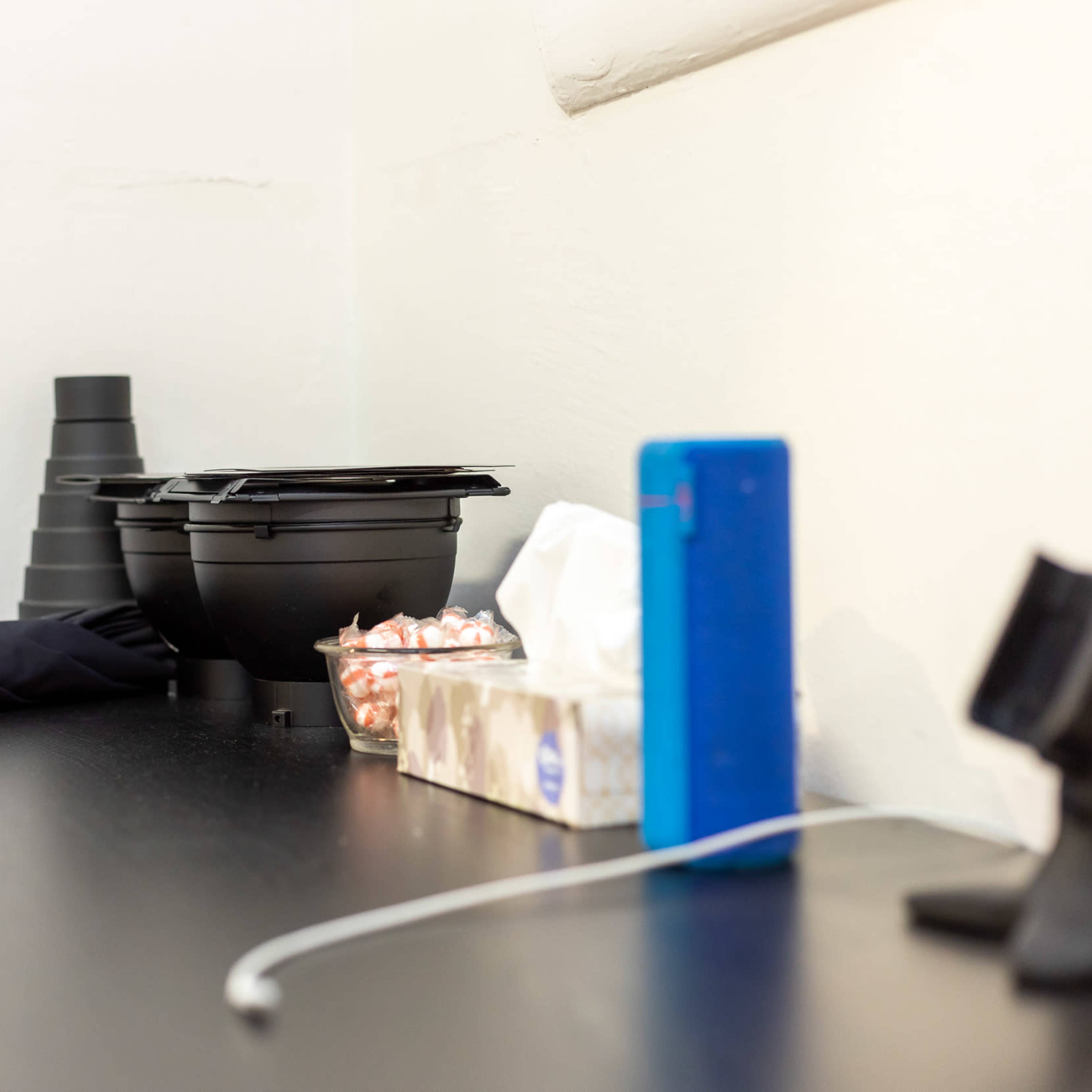 A dark wooden surface holds a black umbrella, two black containers, a bowl of candy, a tissue box, and a blue device with a charging cable.