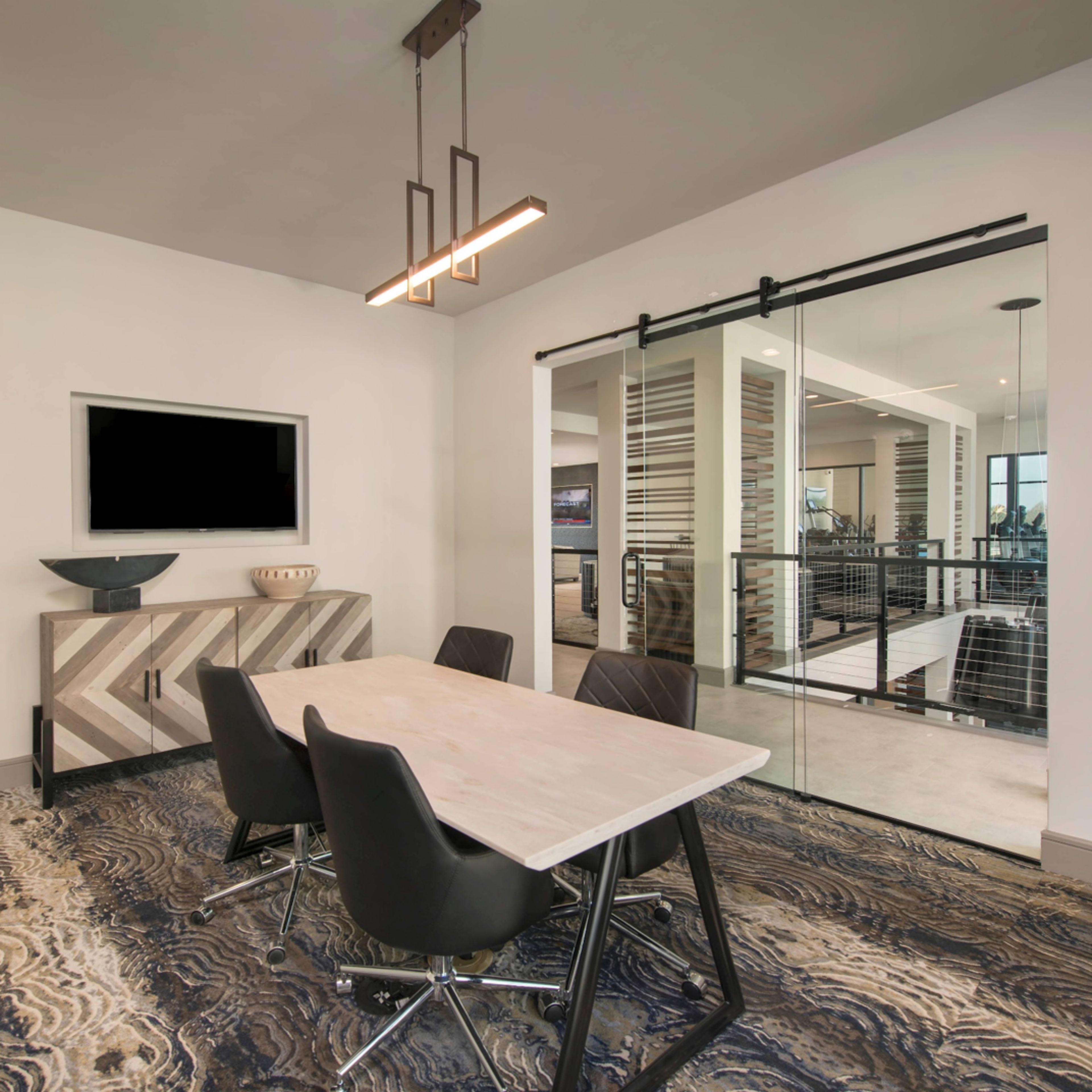 A modern conference room features a rectangular table surrounded by black chairs, with a television mounted on the wall and large glass doors leading to an adjacent space.