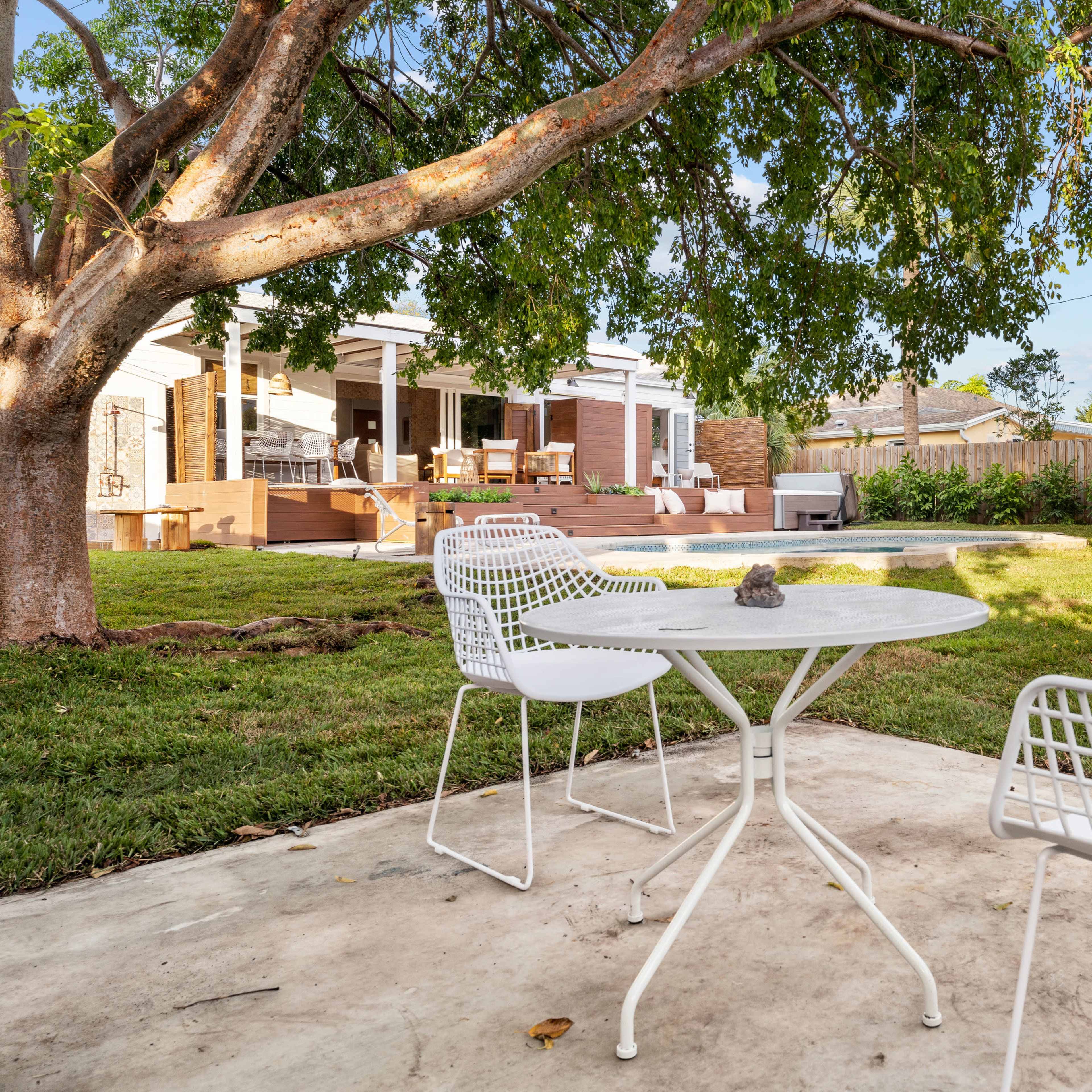 A patio area with three white chairs and a round table is set under a tree, overlooking a backyard with a deck and landscaped greenery.