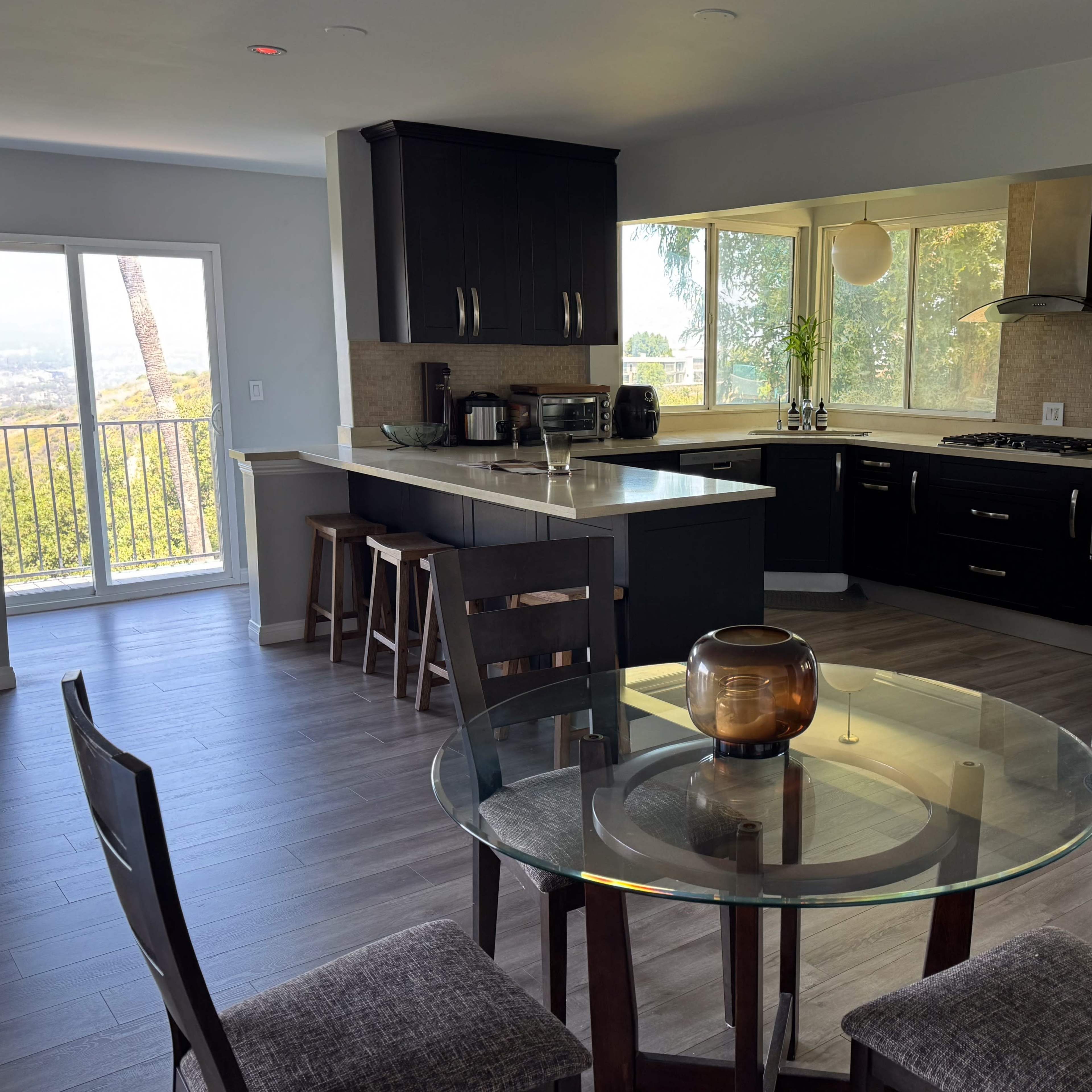 A modern kitchen and dining area with a round glass table, surrounded by wooden chairs, and large windows offering a view of the outdoors.