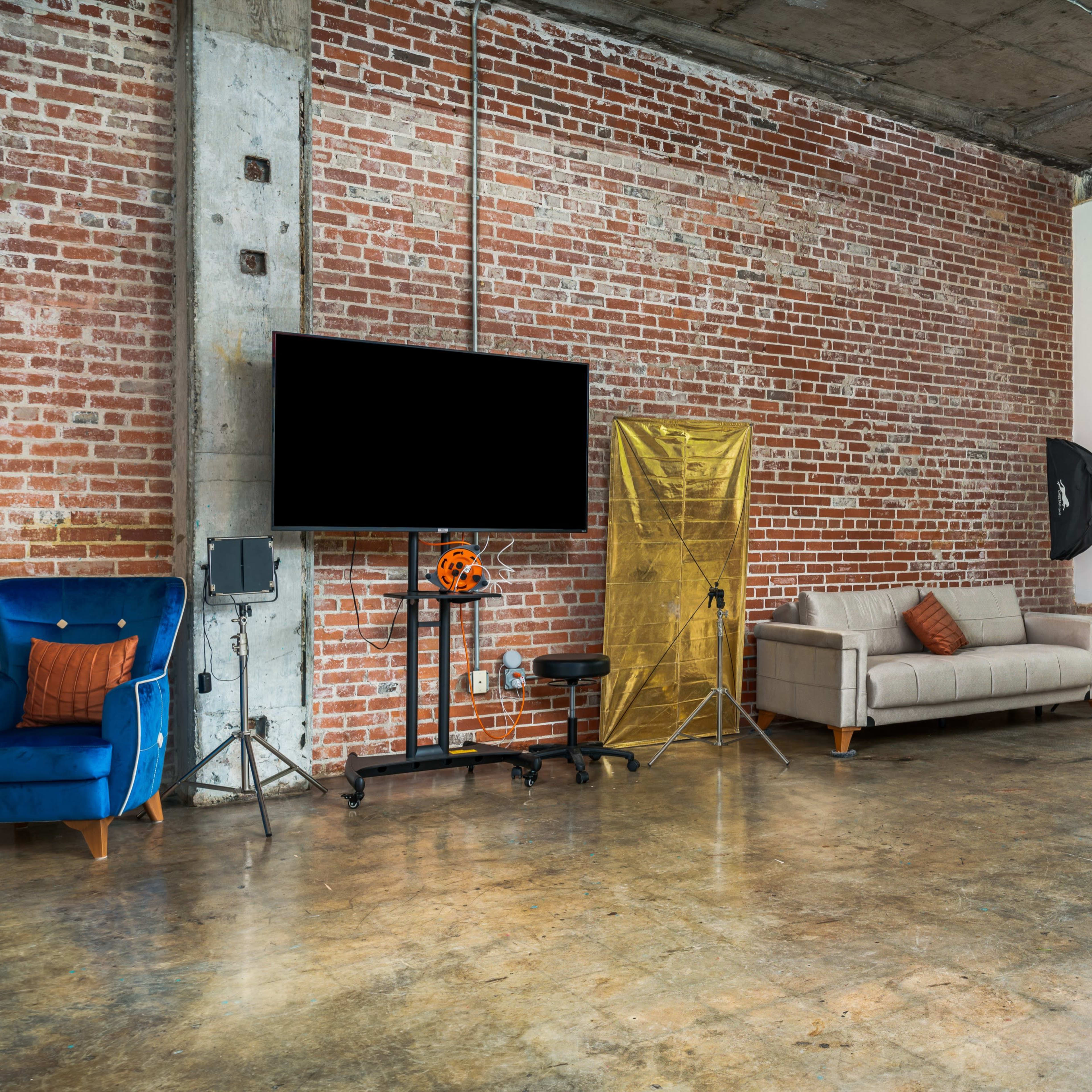 A spacious room with exposed brick walls, a concrete floor, a gray sofa, a television on a stand, and large windows letting in natural light.