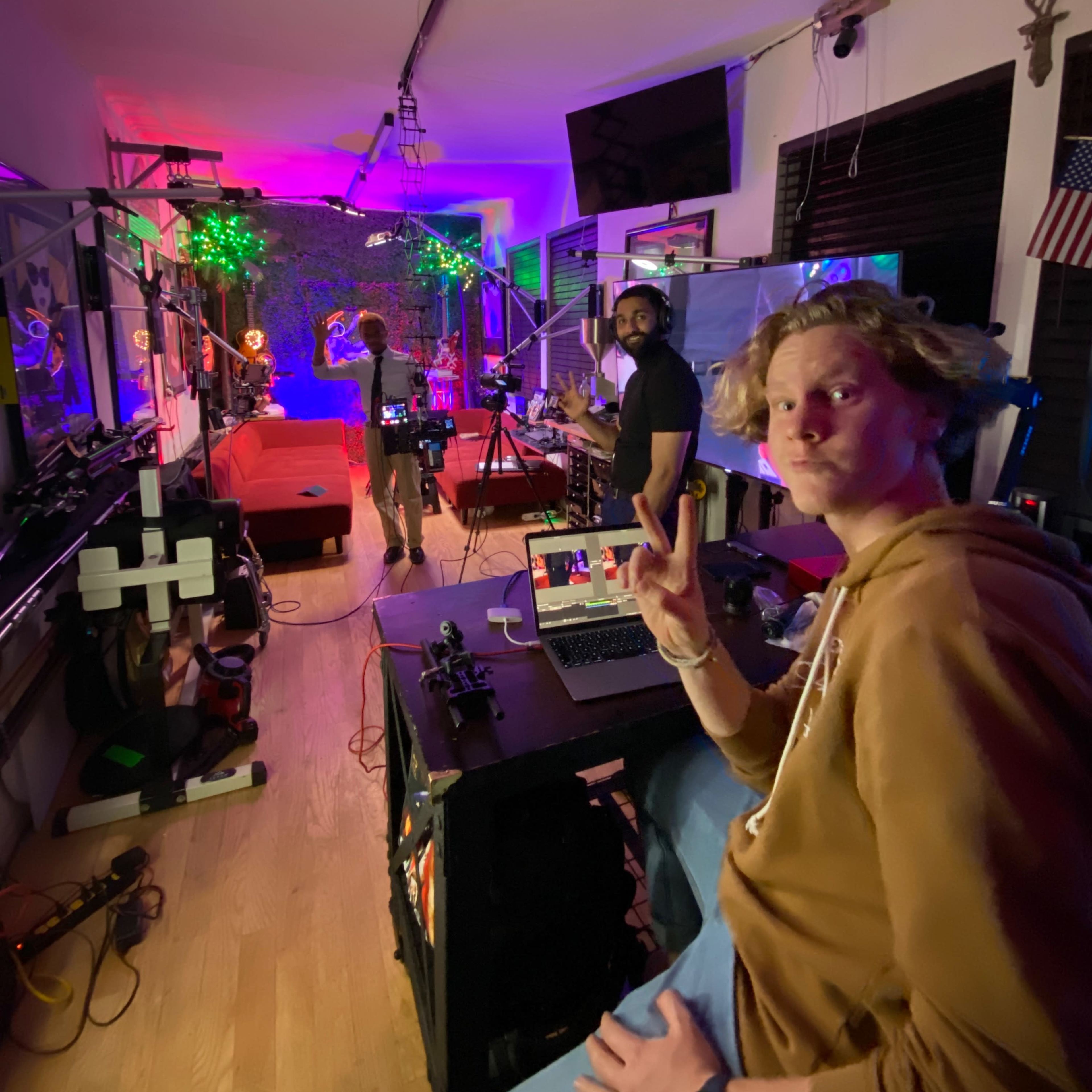 A brightly lit studio space features a person working at a laptop in the foreground, while two others prepare for a shoot in the background surrounded by colorful lighting and equipment.