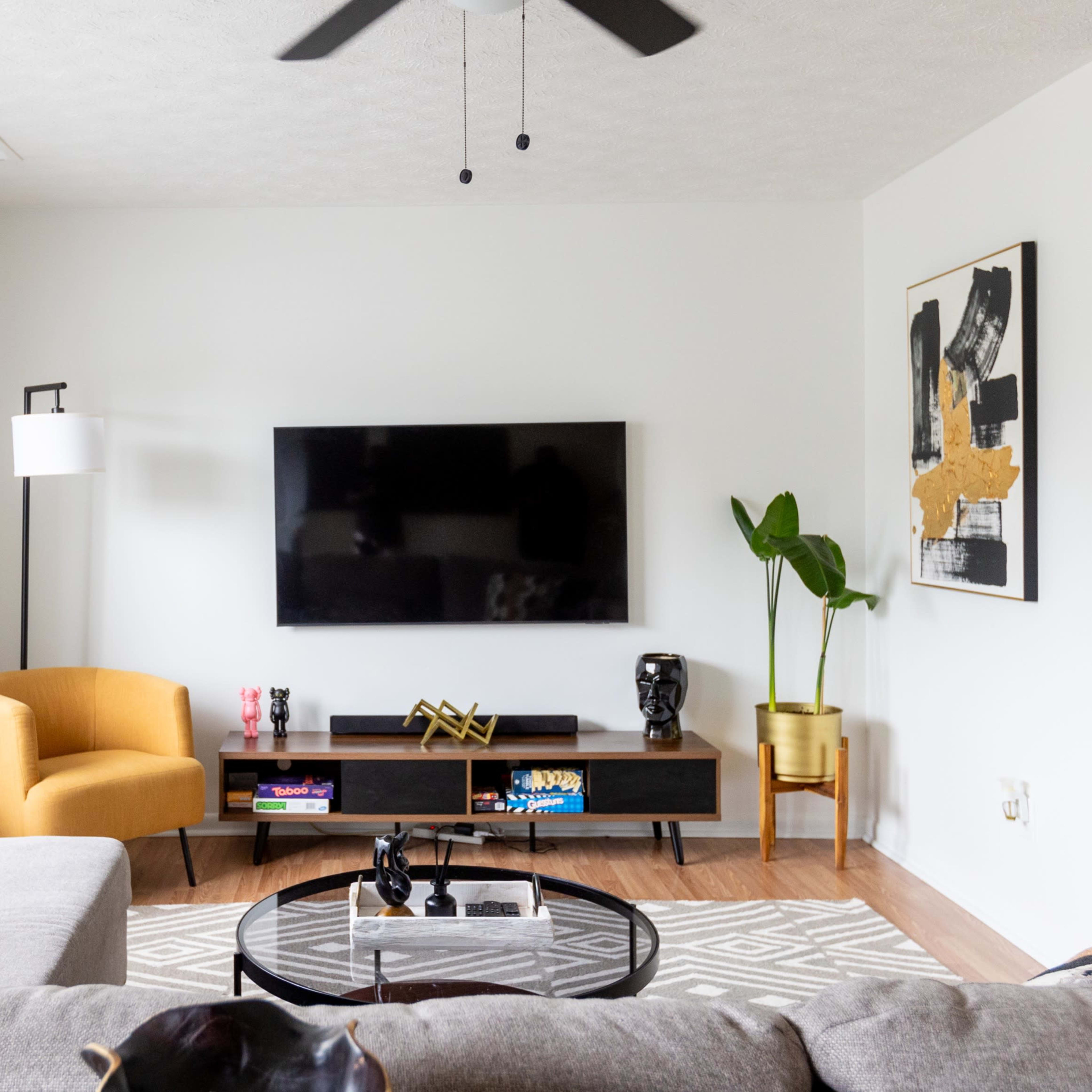 The living room features a gray sectional sofa, a yellow armchair, a black TV mounted on the wall, and a coffee table on a patterned rug.