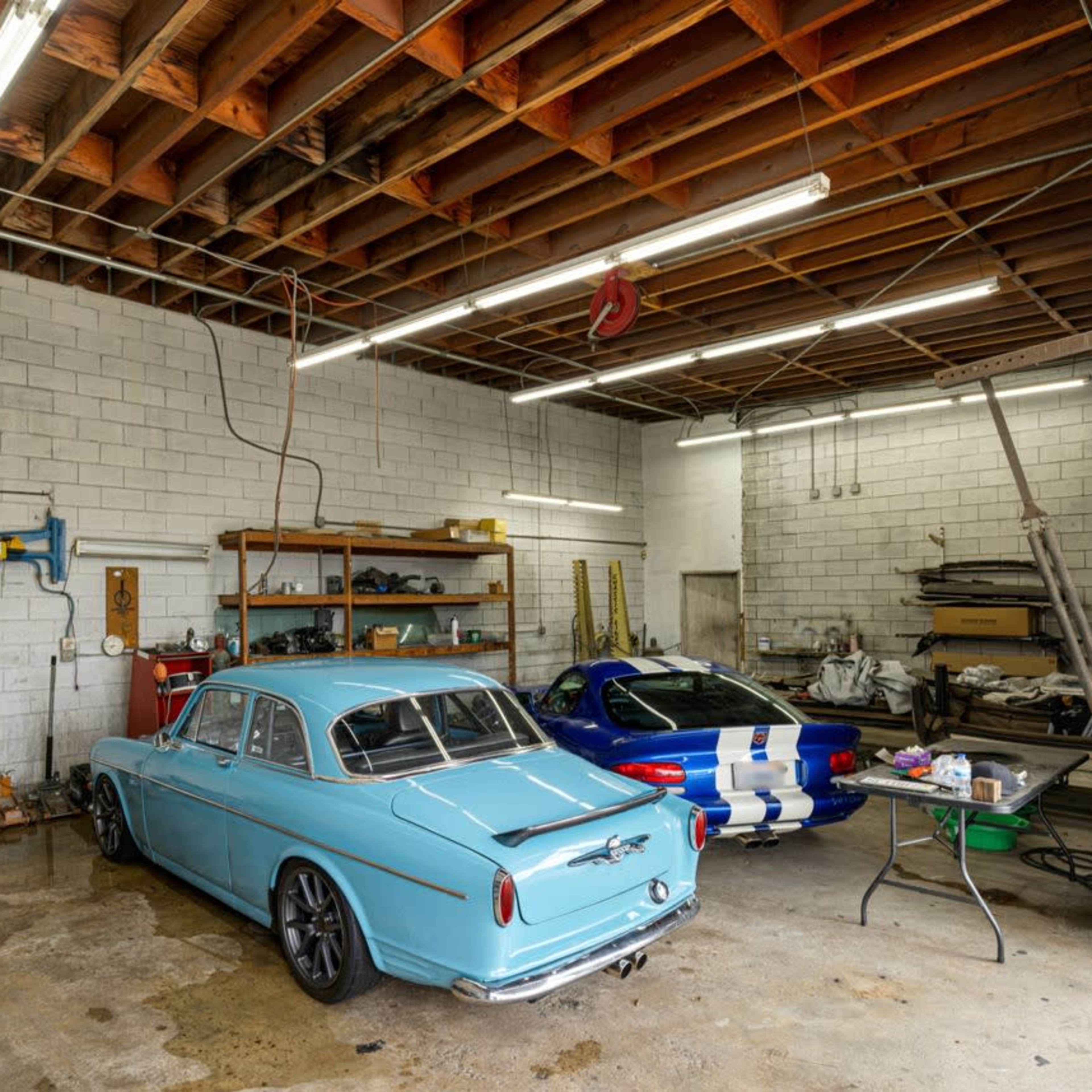 A garage interior with two cars, a light blue sedan and a blue sports car, along with various tools and equipment arranged in the space.