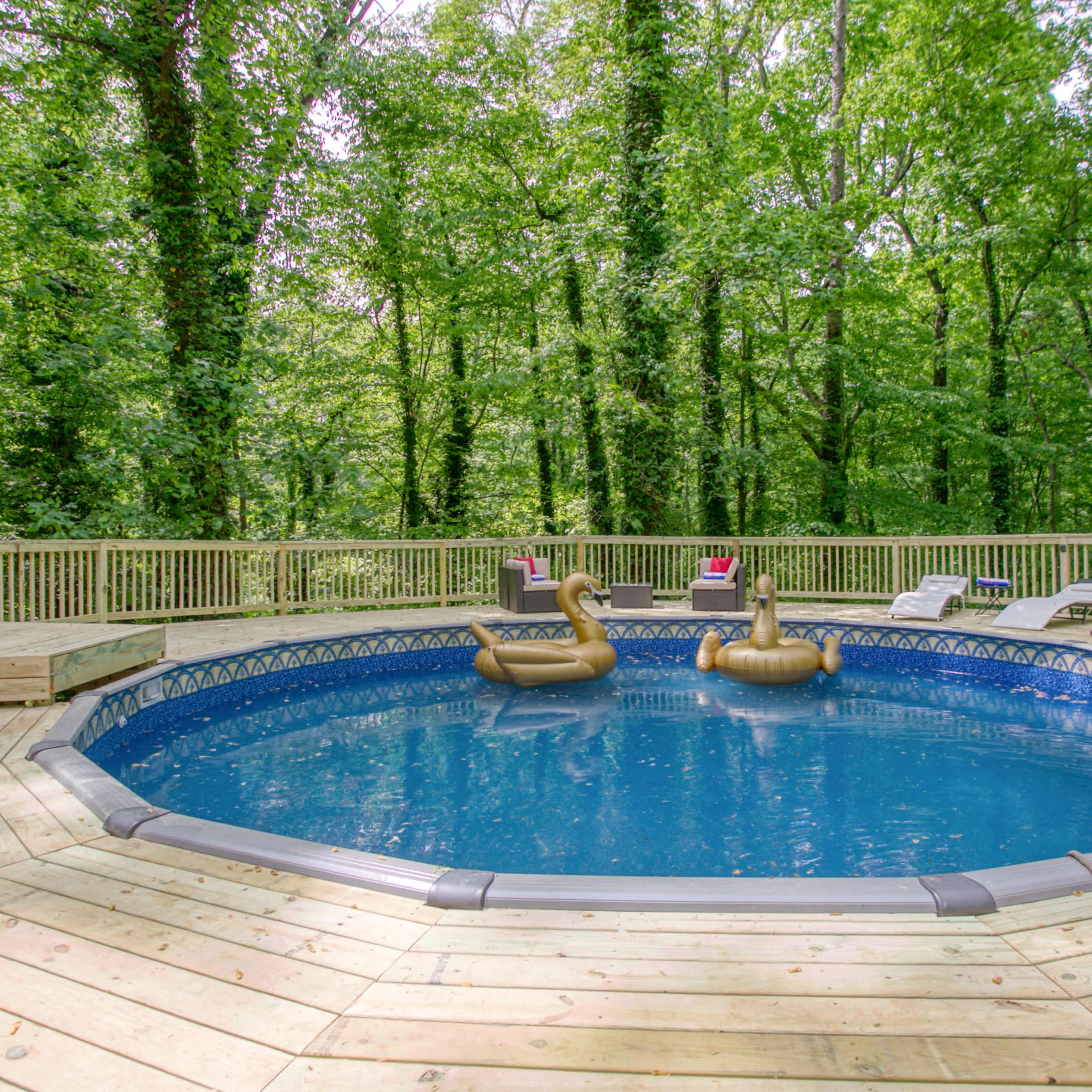 A round above-ground pool with wooden decking is surrounded by lush green trees and lounging chairs.