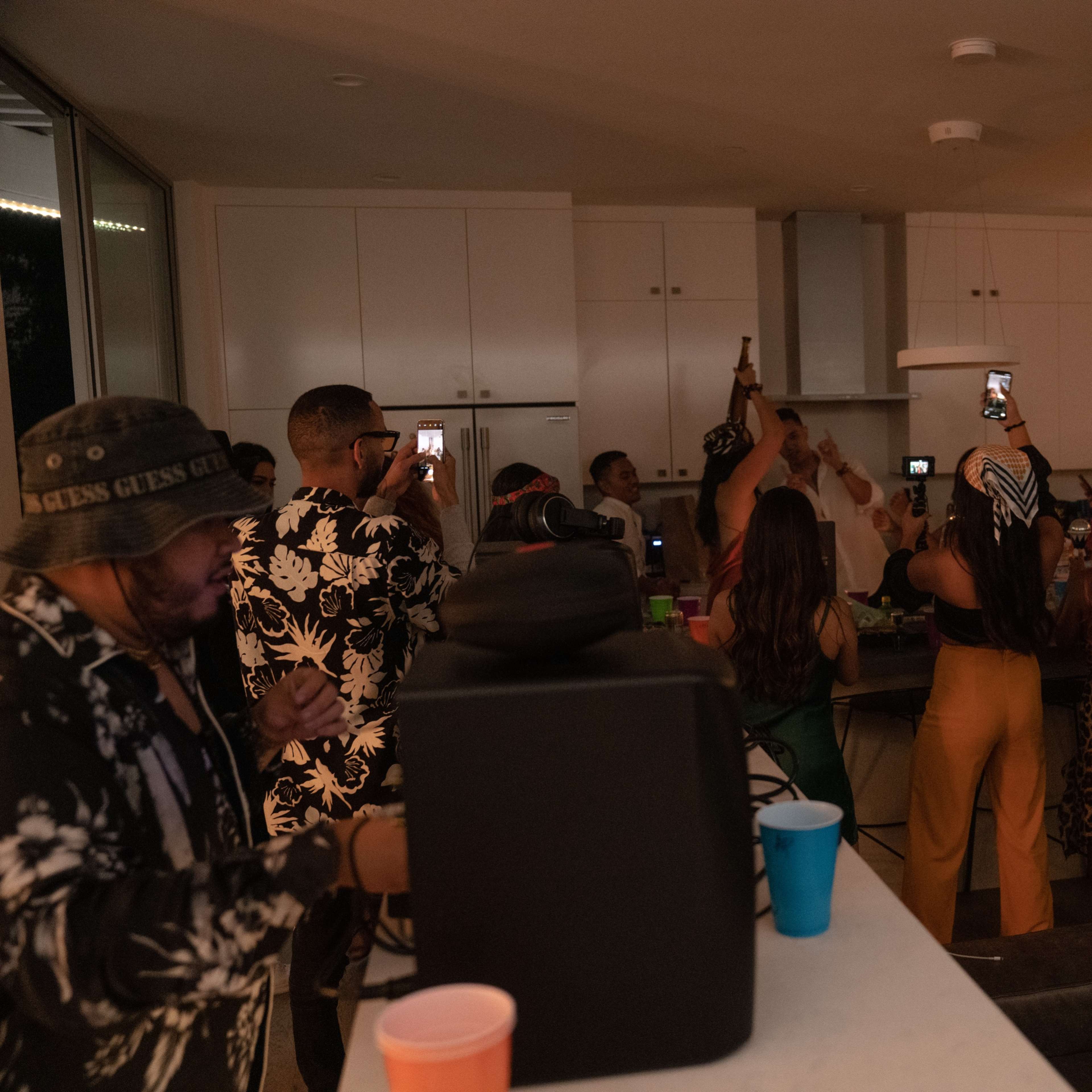 A group of people are gathered in a brightly lit kitchen, taking photos and videos while others perform or celebrate.