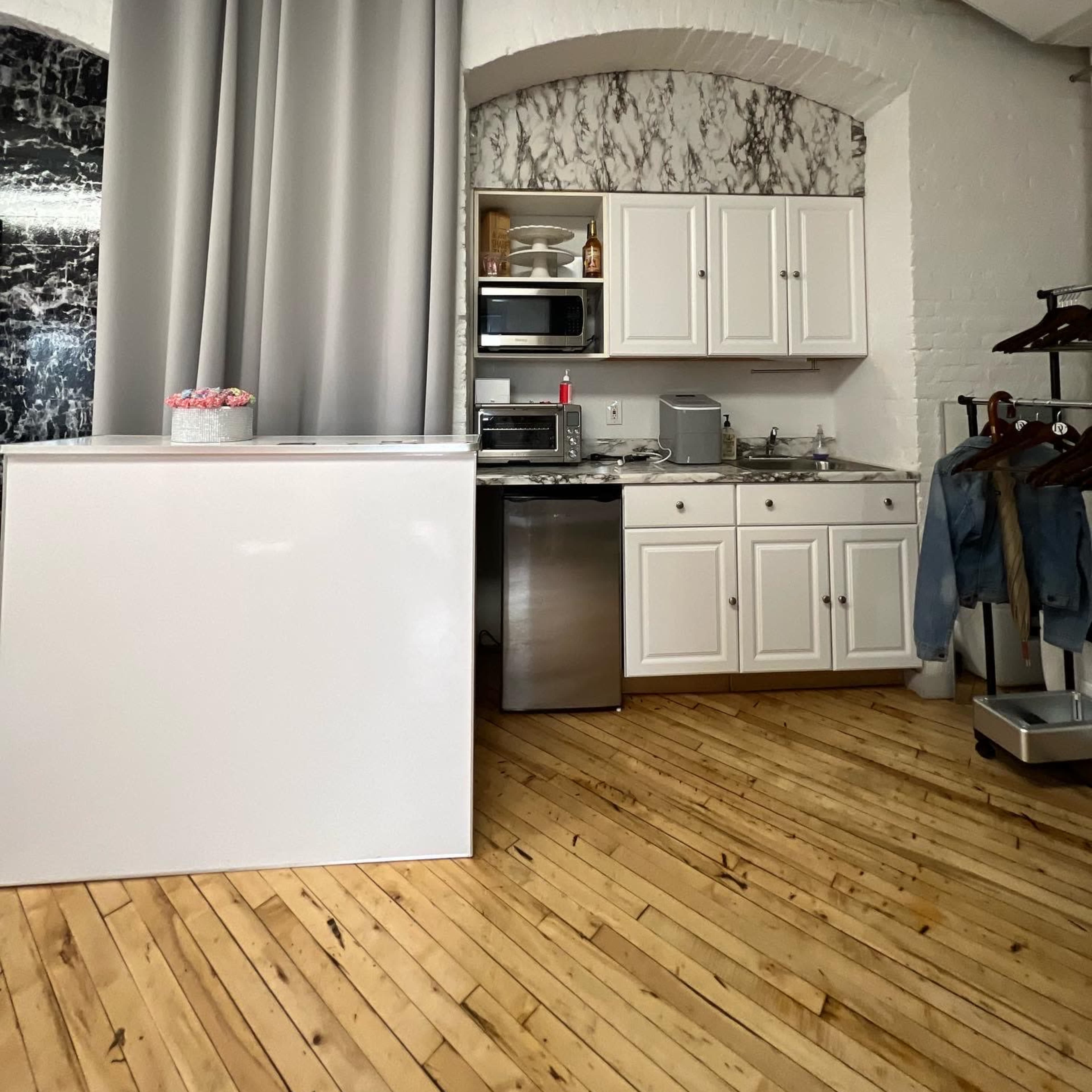 A modern kitchen area with white cabinets and stainless steel appliances, alongside a light wooden floor and gray curtains.