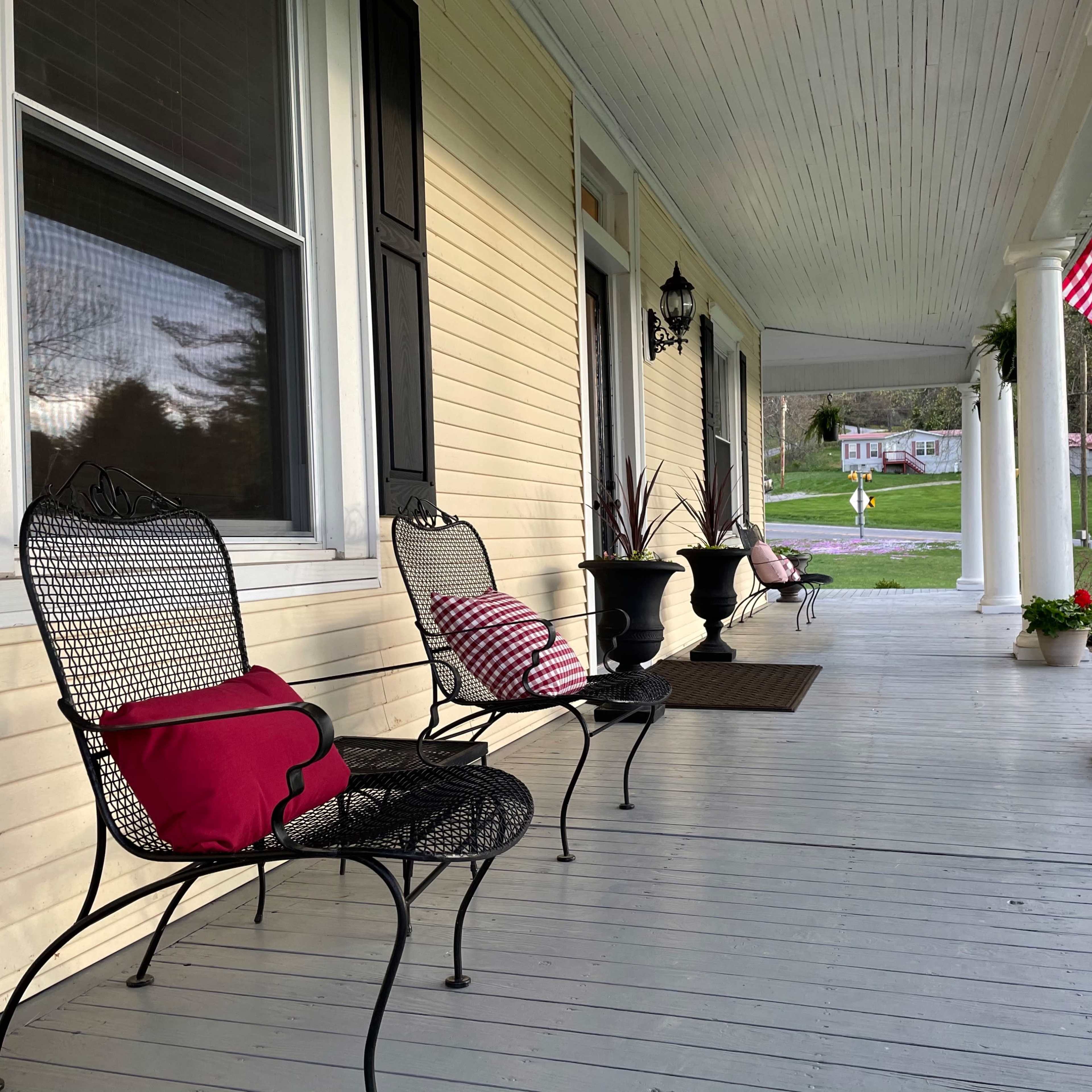 A porch with several black wrought iron chairs and pink cushions, along with potted plants and a view of a grassy area and road in the background.