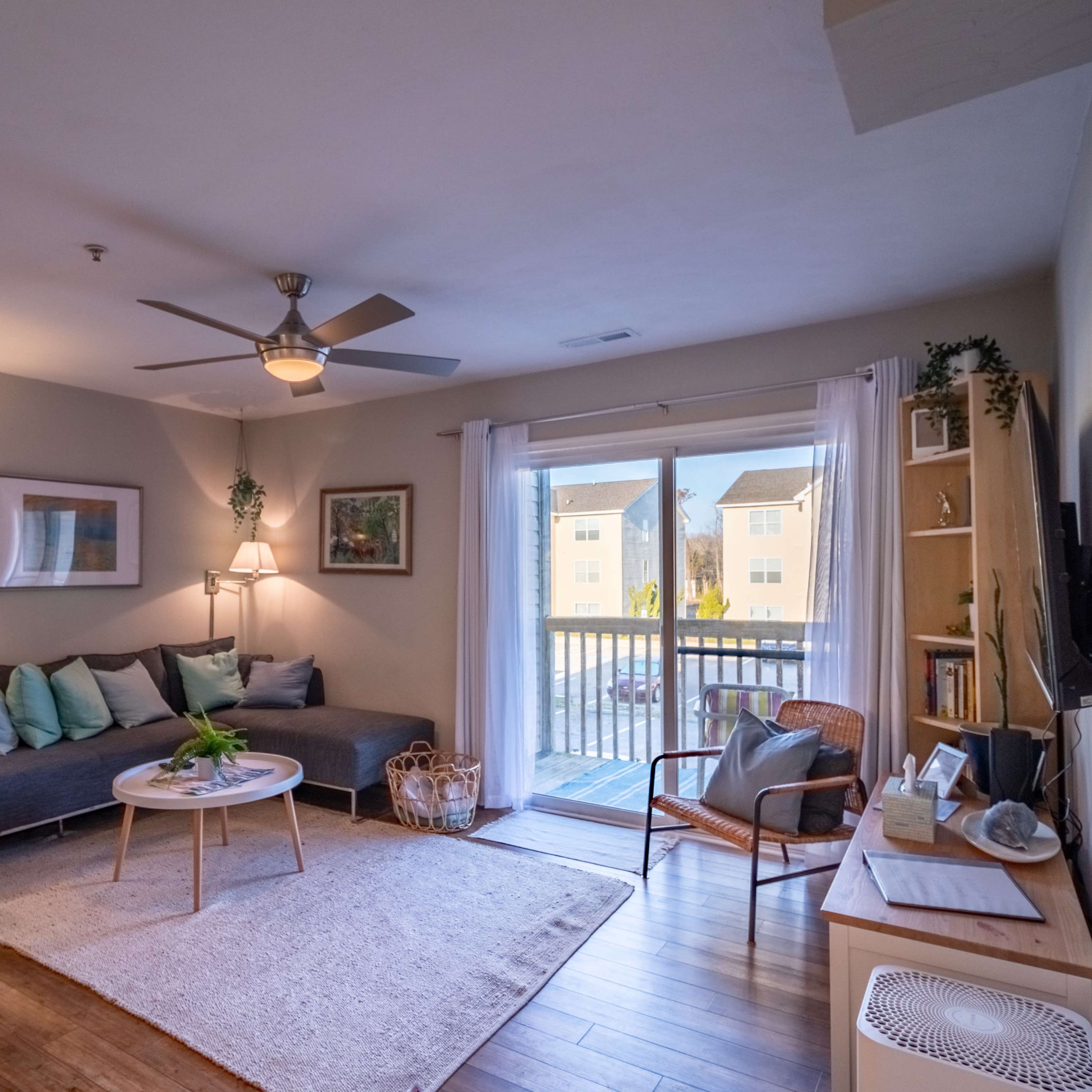 A modern living room features a gray sofa, a round coffee table, and sliding glass doors leading to a patio, with wooden flooring throughout.