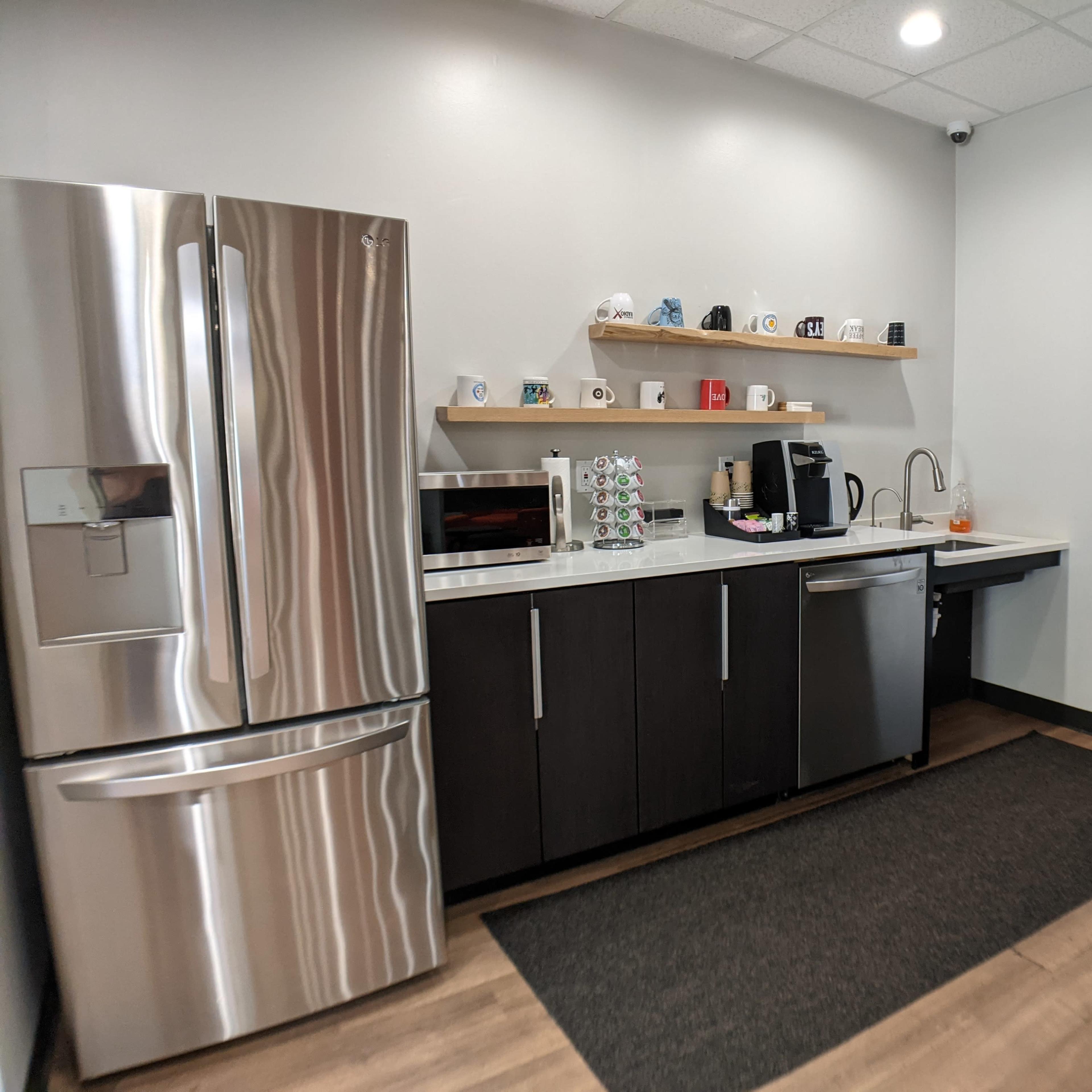 A modern kitchen area with a stainless steel refrigerator, a microwave, a coffee maker, and a sink, arranged along a dark cabinetry wall with shelves displaying mugs.