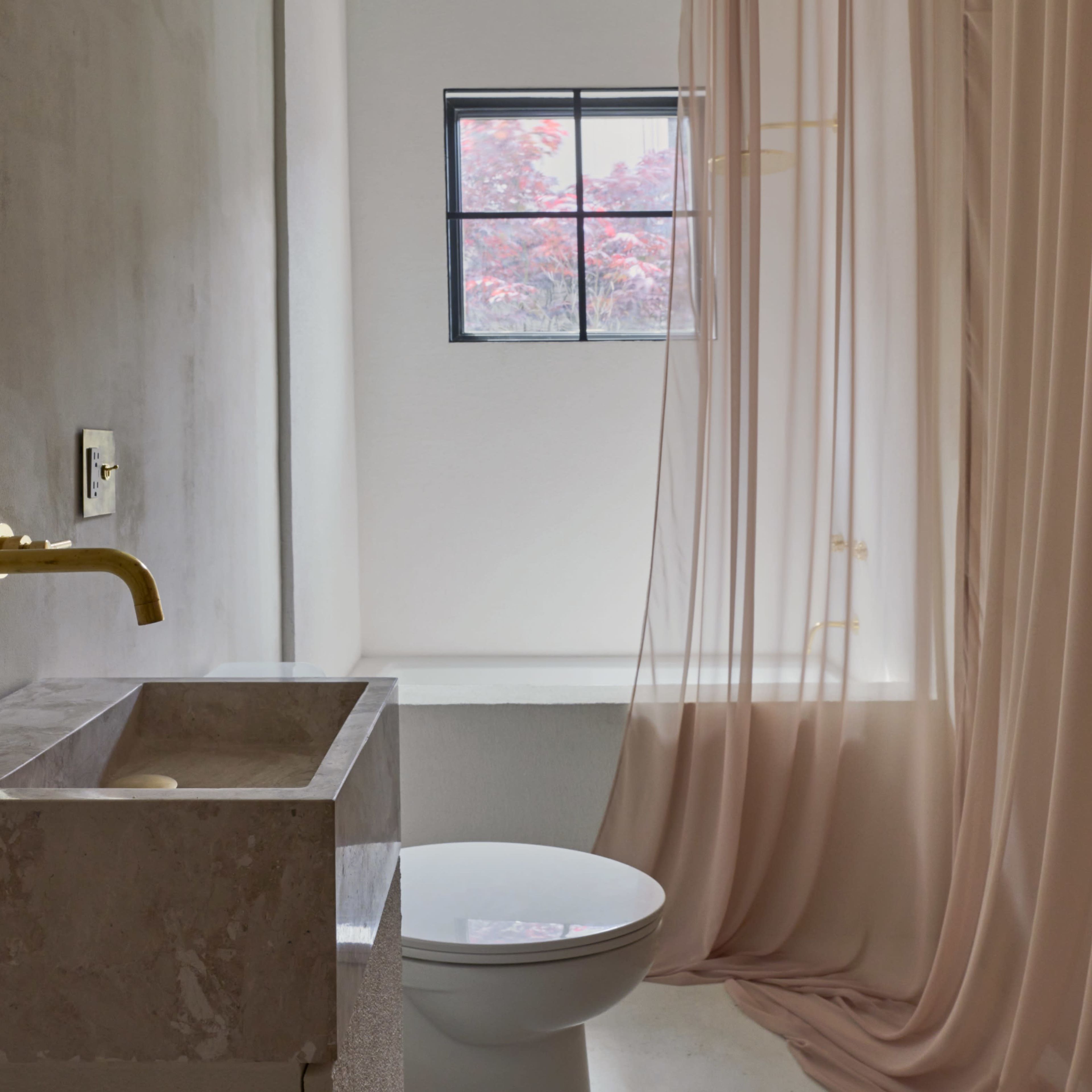 A modern bathroom featuring a gold faucet, a marble sink, a toilet, and a sheer curtain separating the space from a window with a view of red foliage outside.