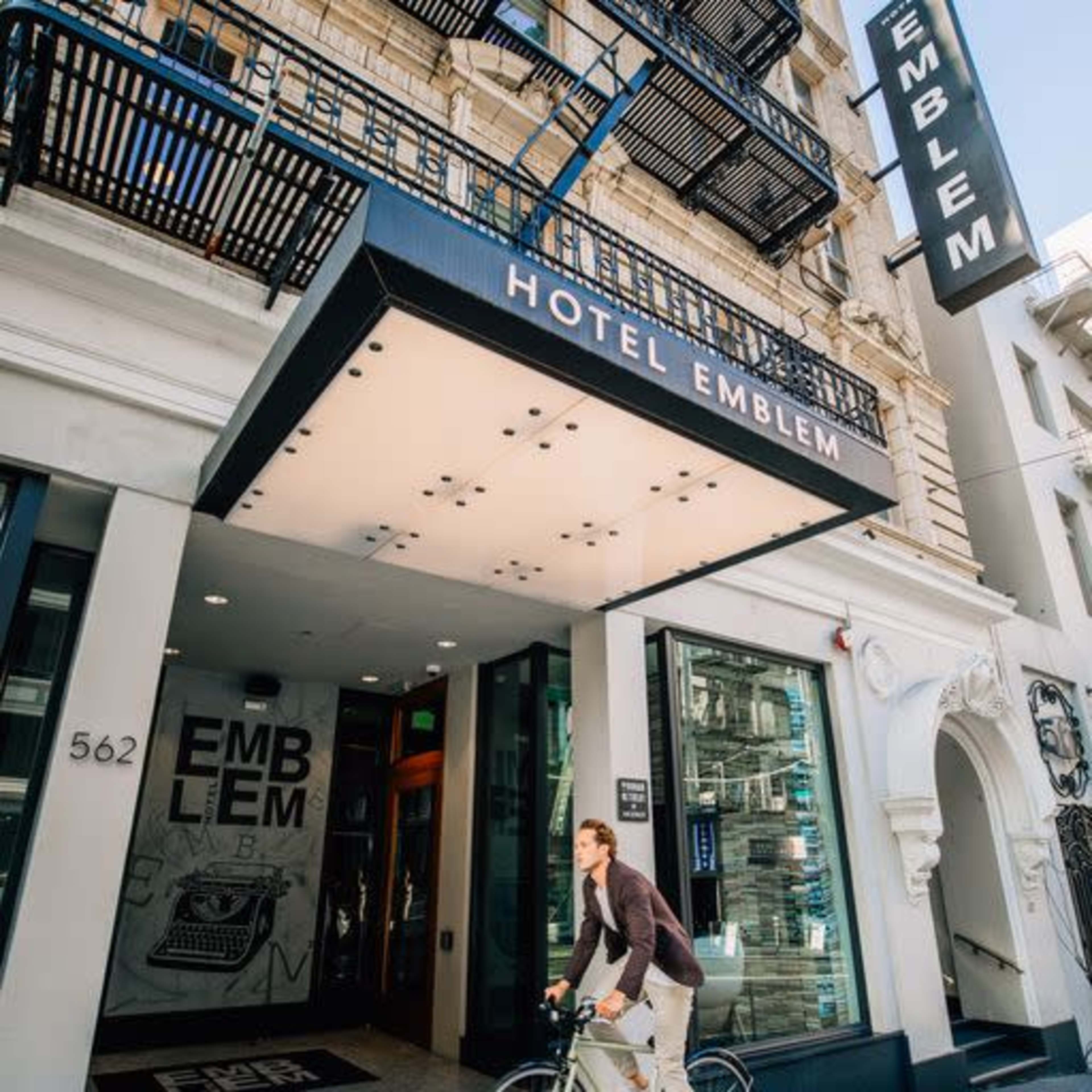A man rides a bicycle past the entrance of Hotel Emblem, showcasing its modern design and signage.
