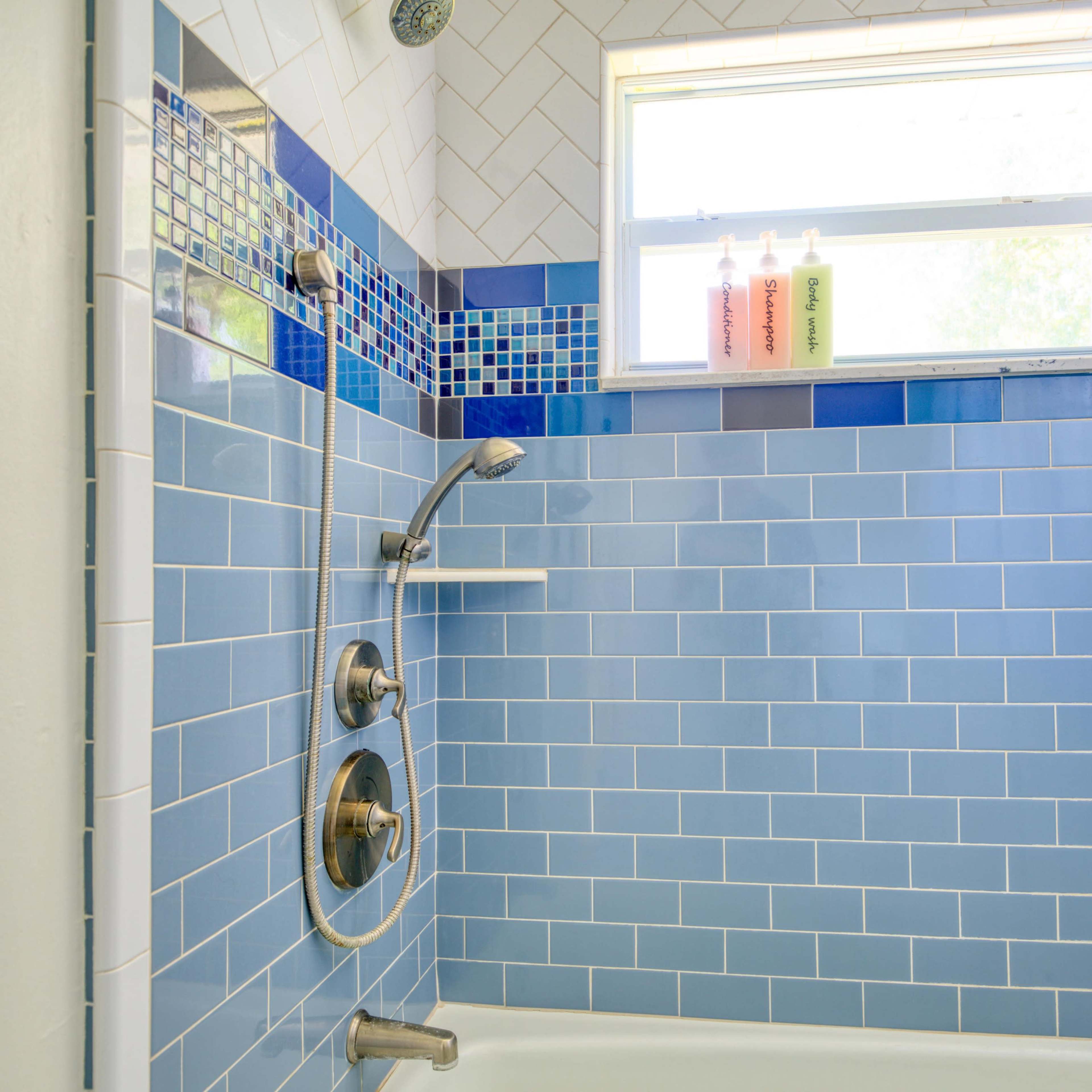 A tiled shower area with blue tiles, a showerhead, a faucet, and three bottles on the windowsill.