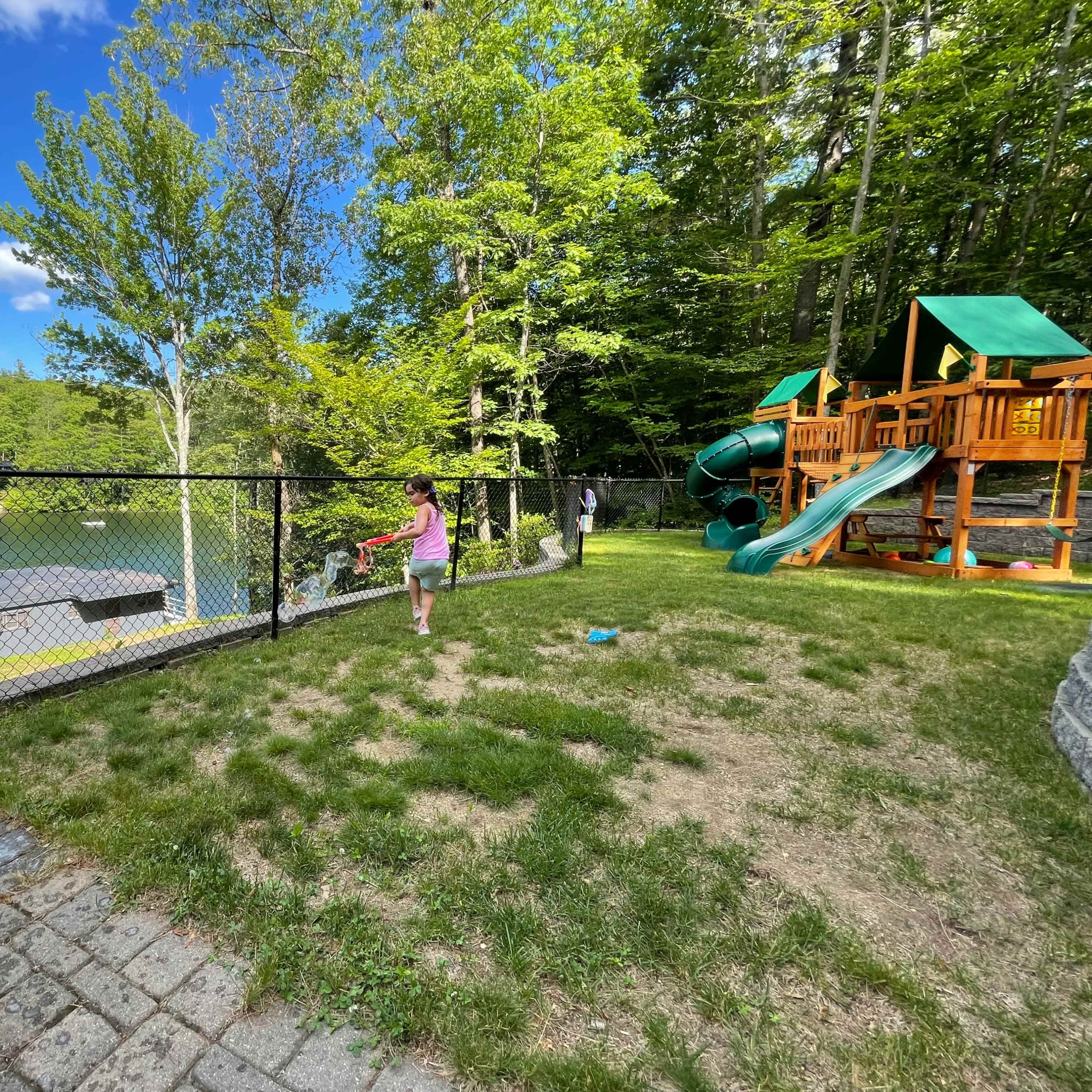 A child plays near a fenced backyard by a lake, with a playground structure visible in the background.
