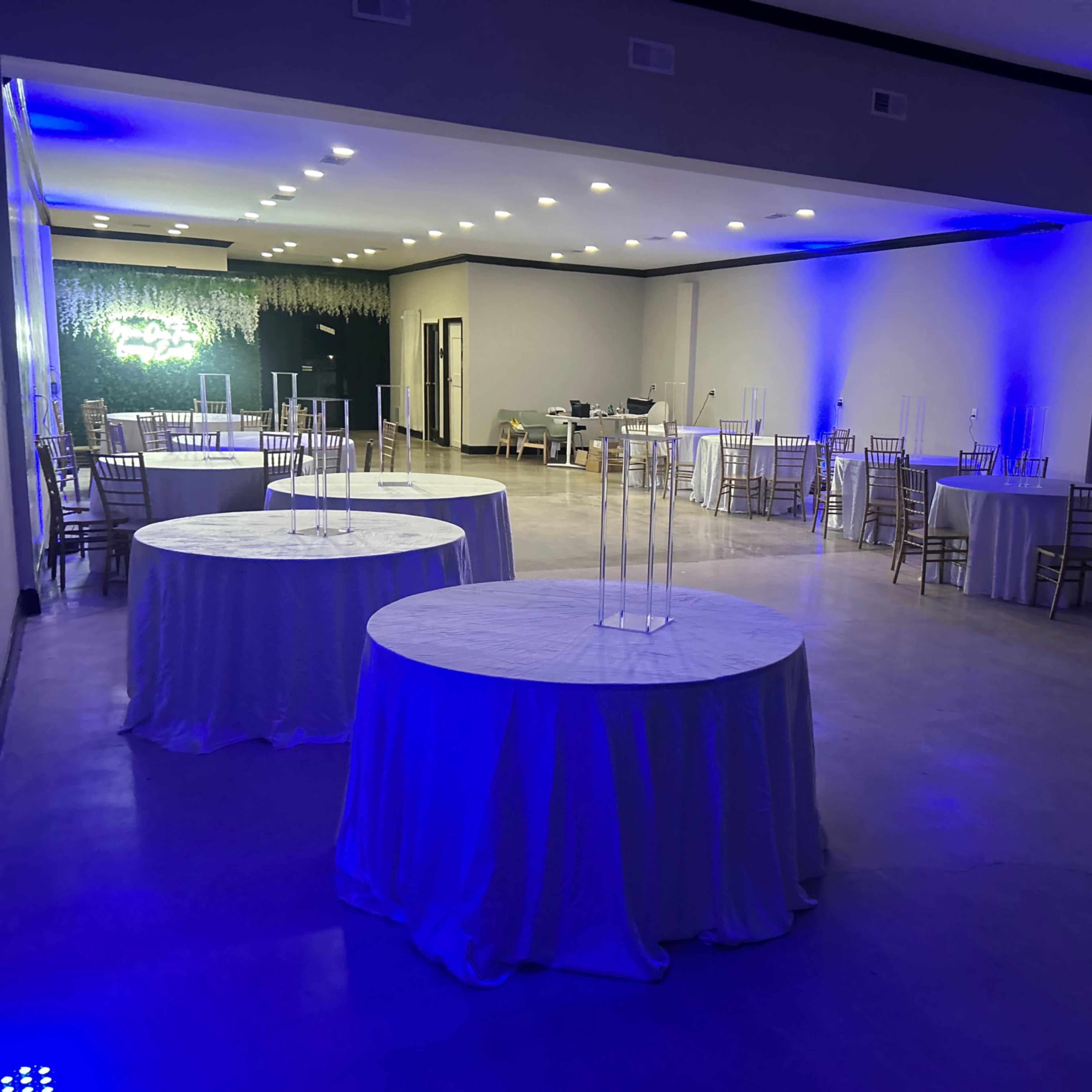 A spacious event venue with several round tables covered in white tablecloths and illuminated by blue lighting.