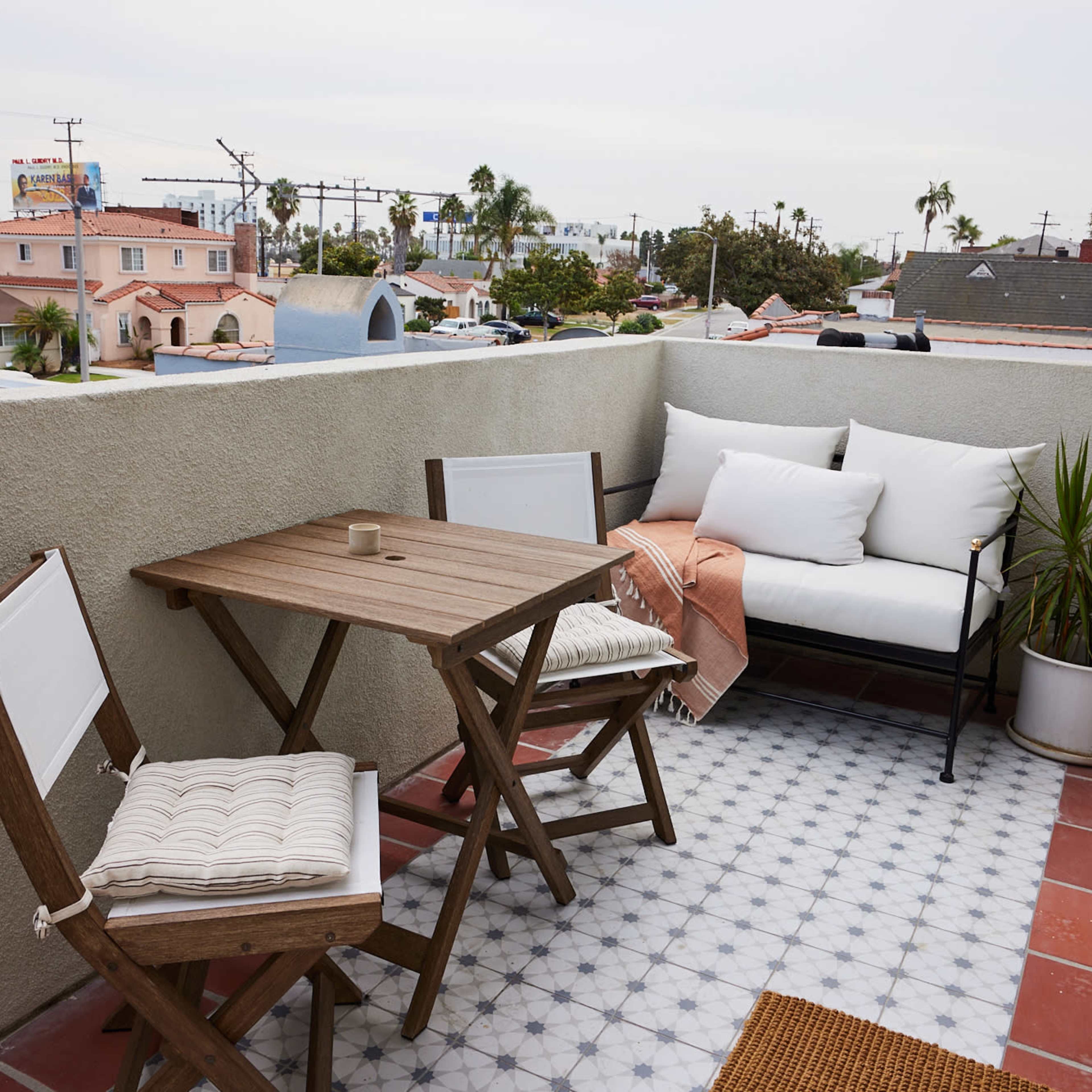 A small balcony with a wooden table and two chairs, alongside a sofa and a potted plant, overlooking a neighborhood with scattered rooftops.