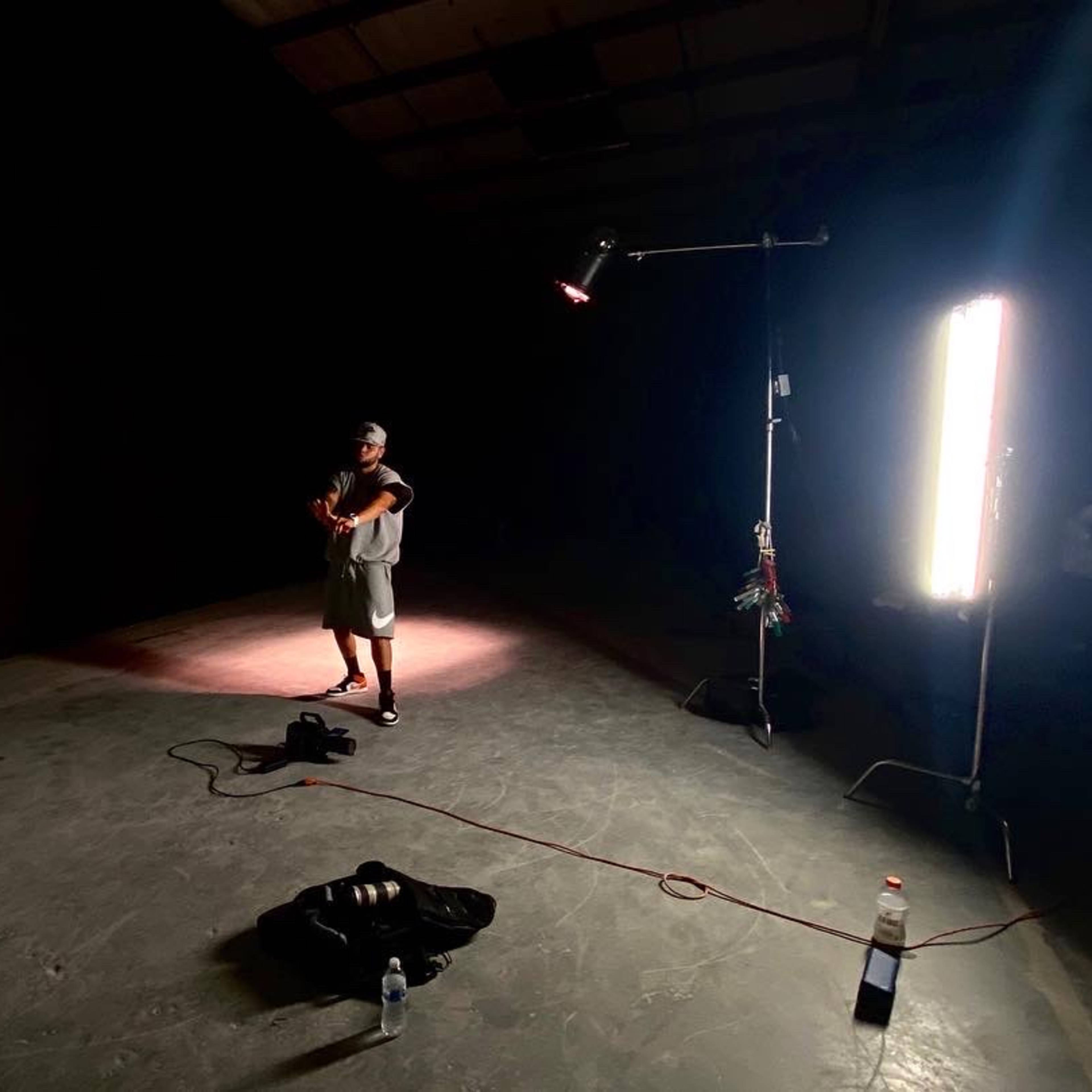 A person stands in a dimly lit studio with a violin, near a white panel and some lighting equipment.