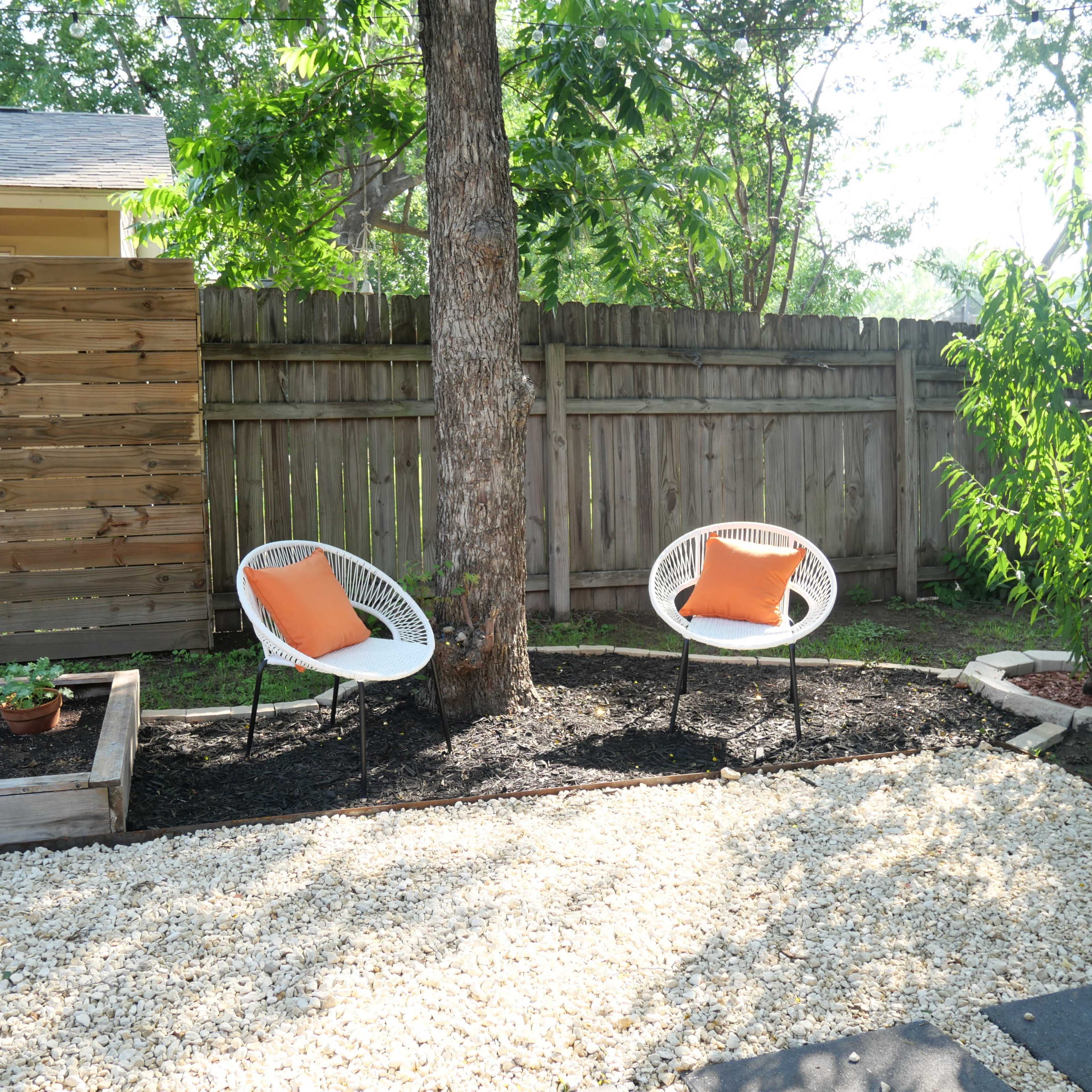 Two white chairs with orange cushions are placed under a tree in a gravel area surrounded by a wooden fence and a small garden bed.
