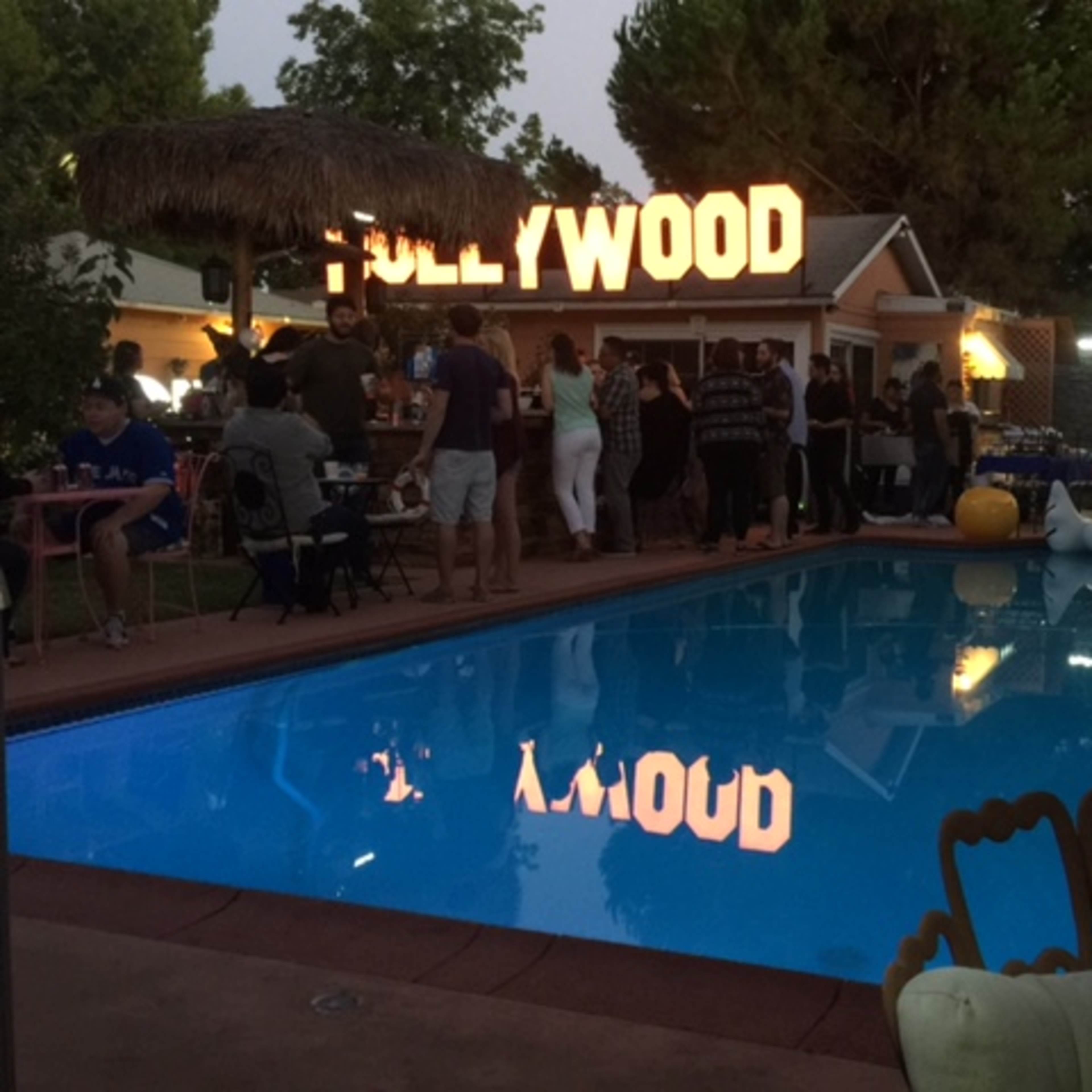 A lively gathering takes place around a pool, with a large "HOLLYWOOD" sign illuminated in the background.