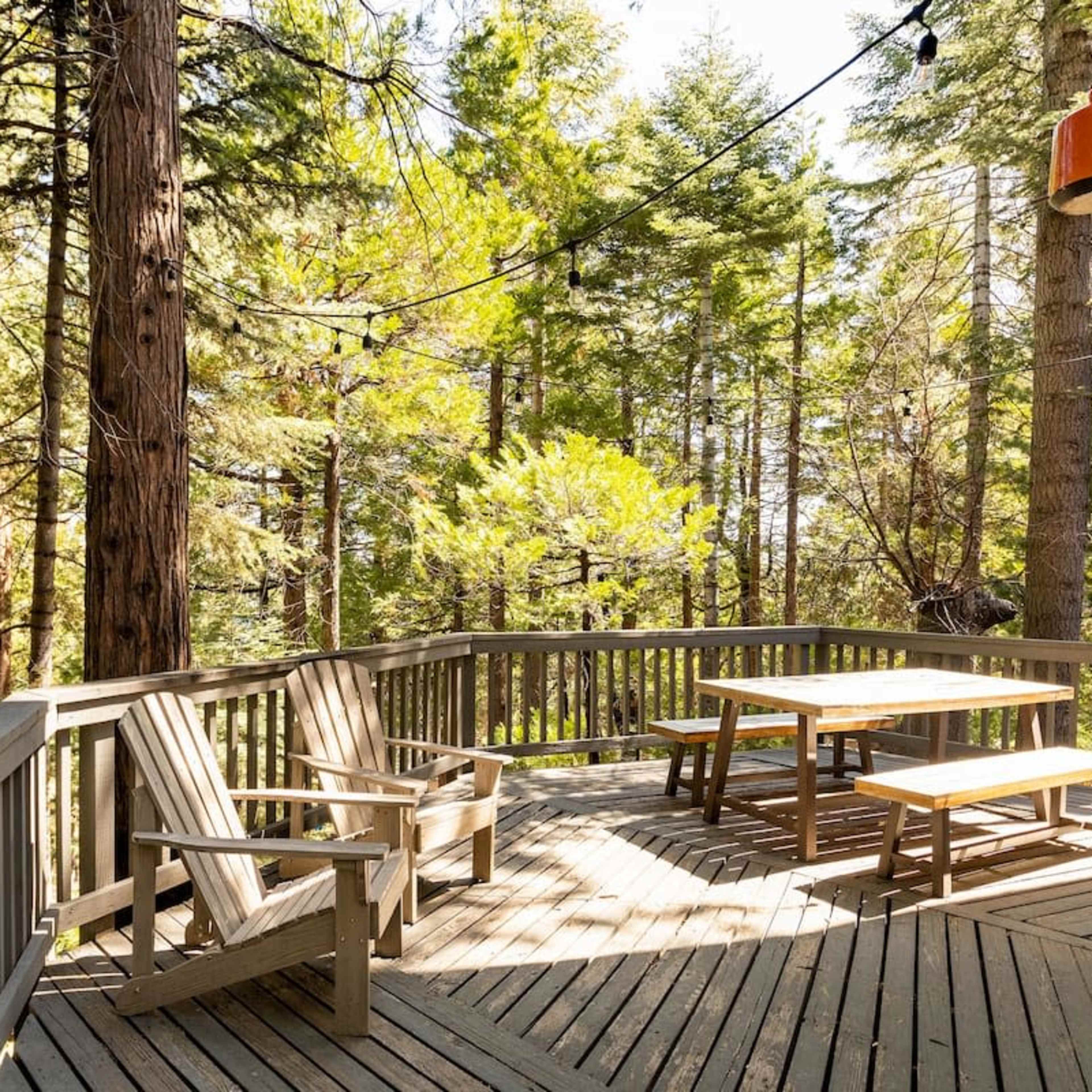 A wooden deck with two chairs and a picnic table is surrounded by tall trees in a forested area.