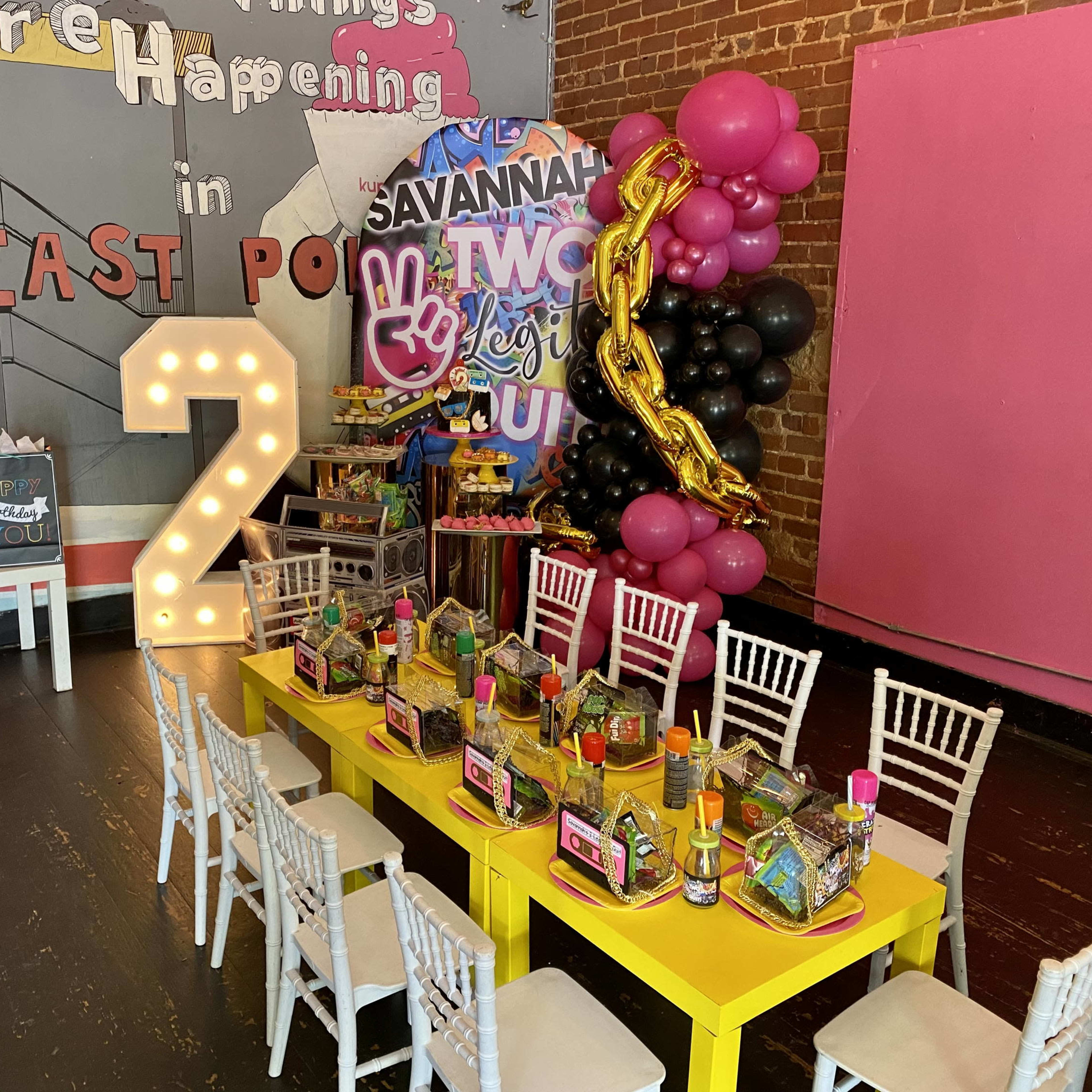 A festive party setting features a yellow table arranged with colorful party favors and decorations, including a large pink and black balloon display and a prominent "2" marquee light, set against a brick wall.