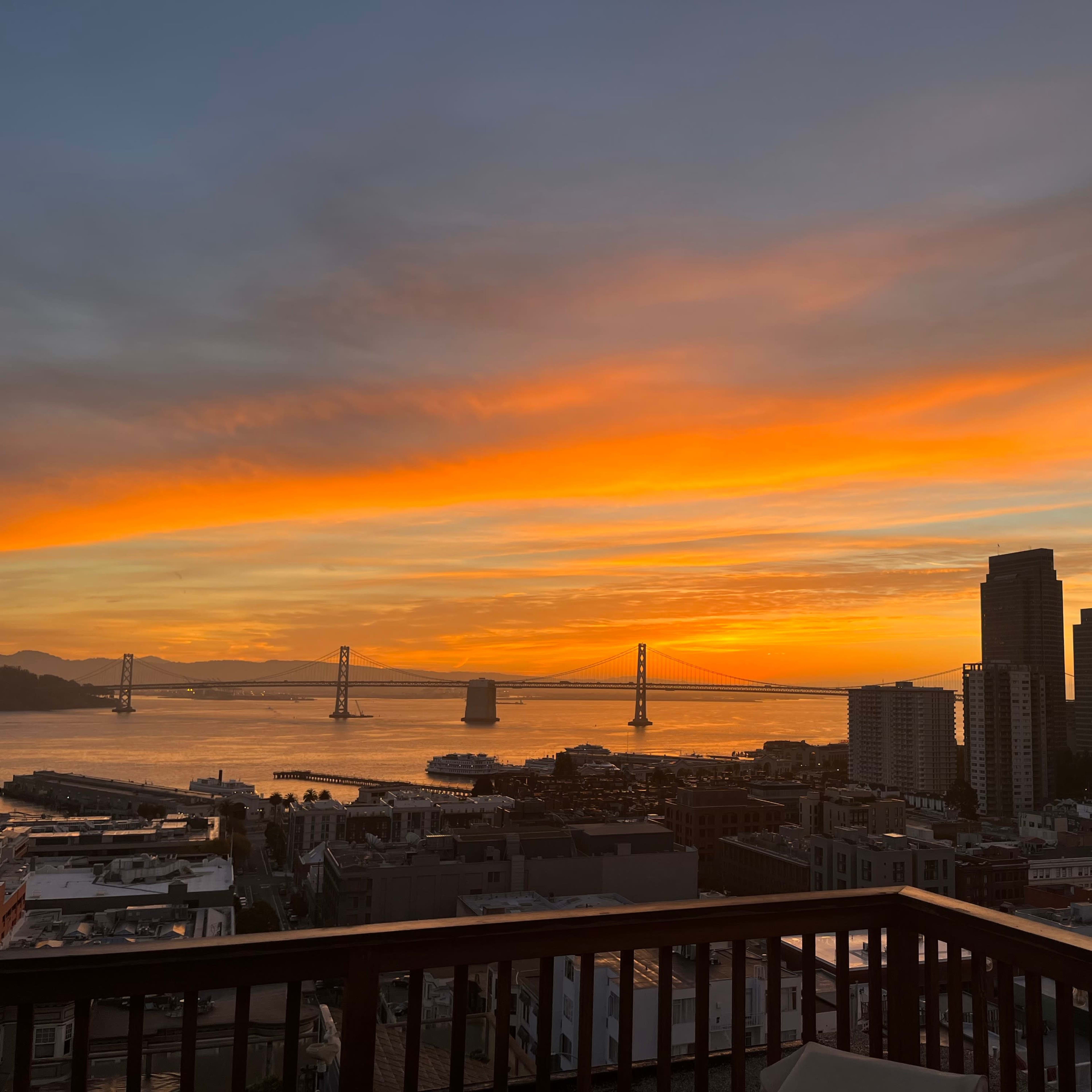 A vibrant sunset over the San Francisco Bay, with the Bay Bridge visible and the skyline of buildings in the foreground.
