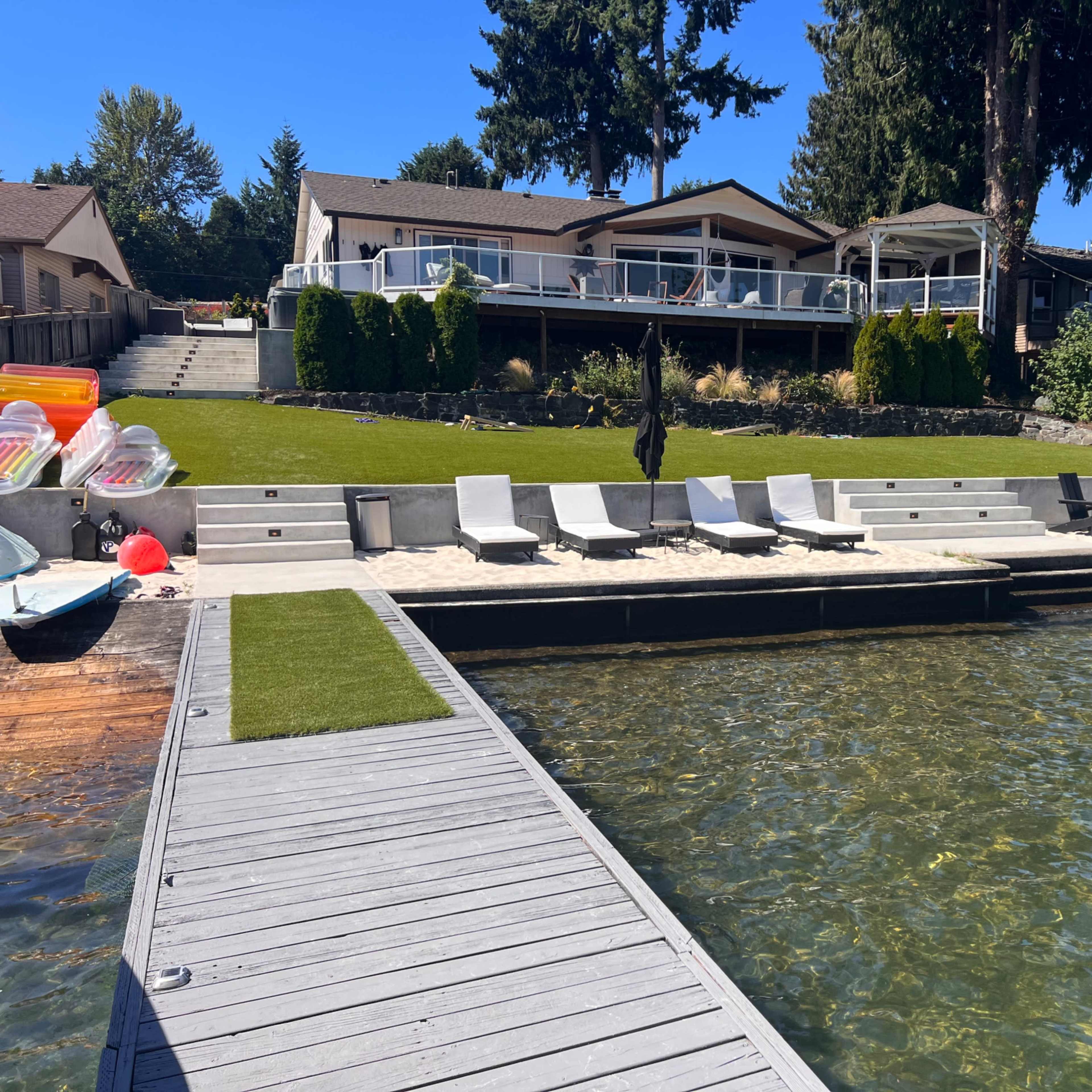 A wooden dock extends over water, leading to a sandy area with loungers, surrounded by a landscaped lawn and waterfront homes.