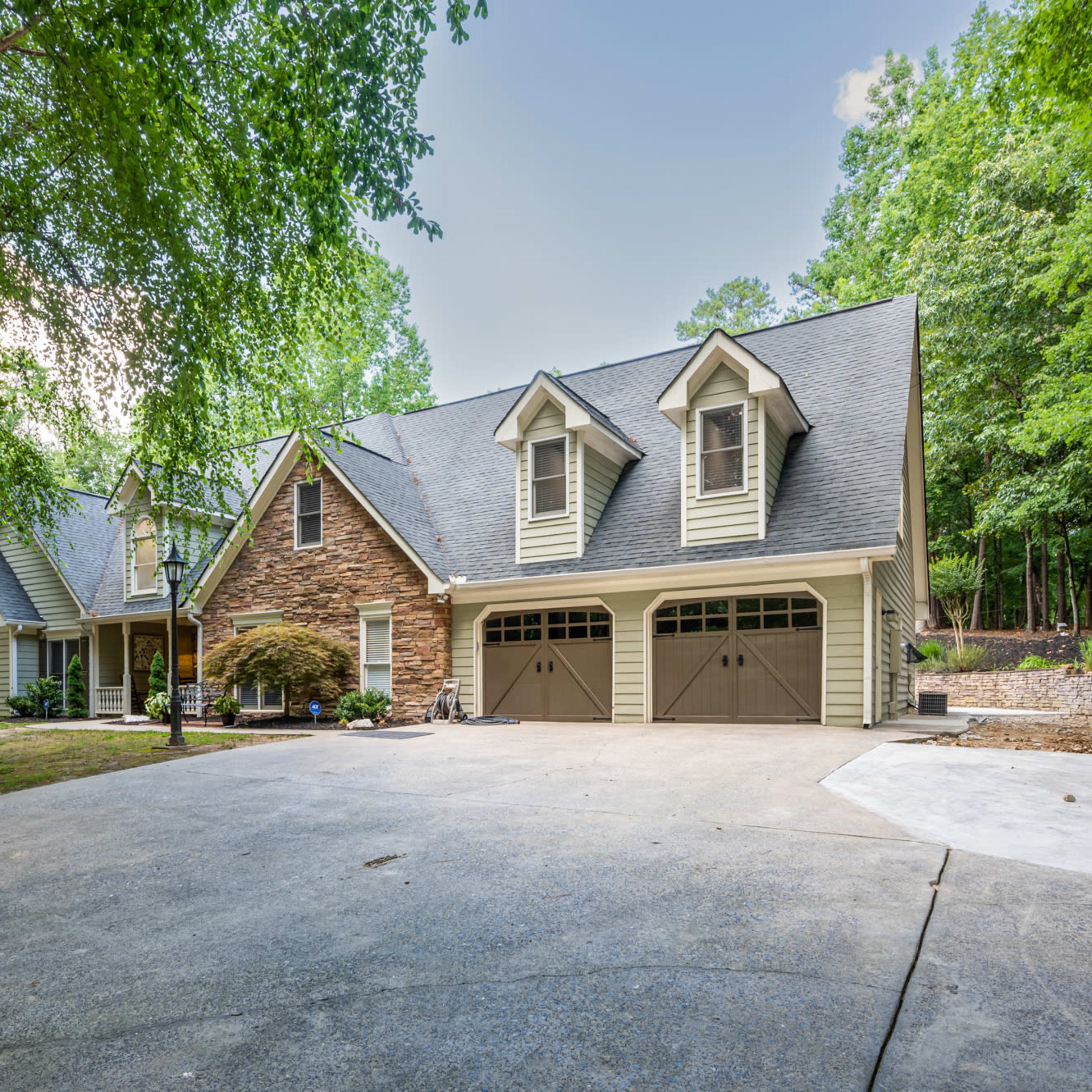 A large, two-story house with a stone and wooden facade is situated under trees, featuring a two-car garage and a concrete driveway.
