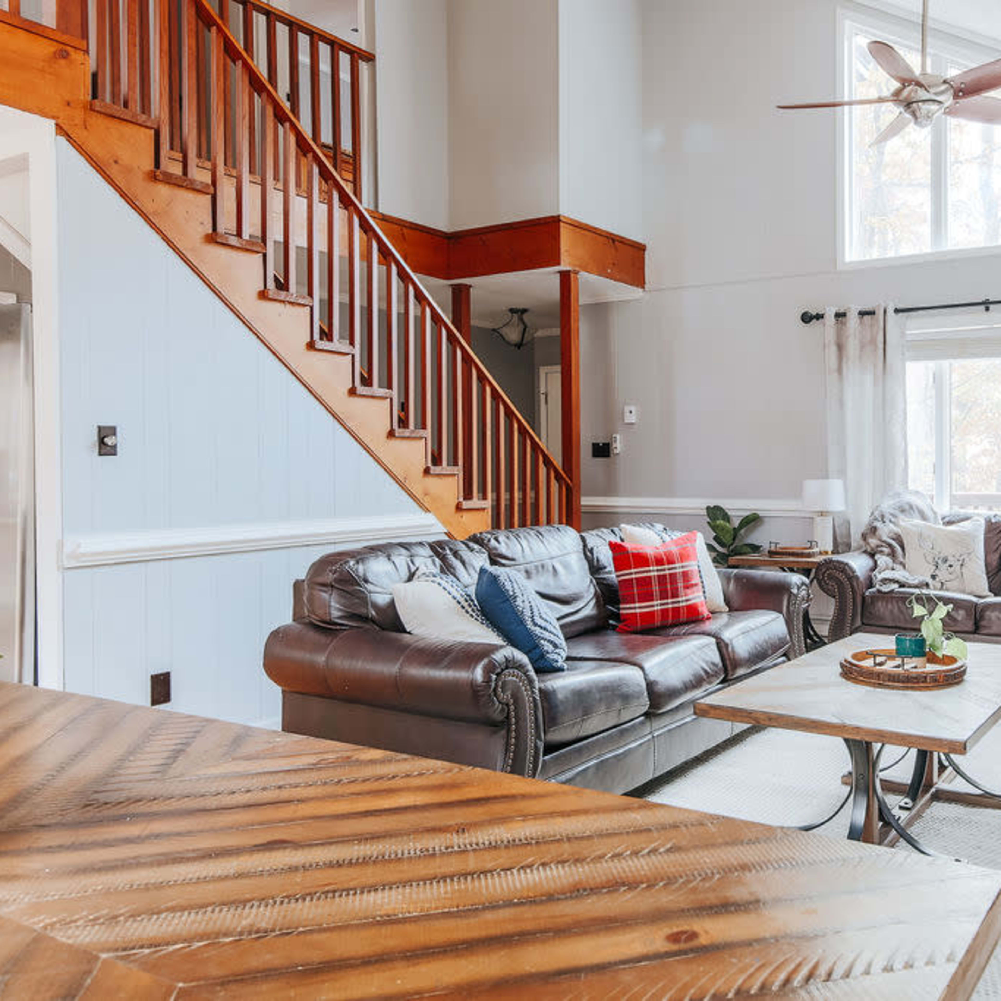 A cozy living room with a leather sofa and armchair, a wooden coffee table, and large windows allowing natural light to enter the space.