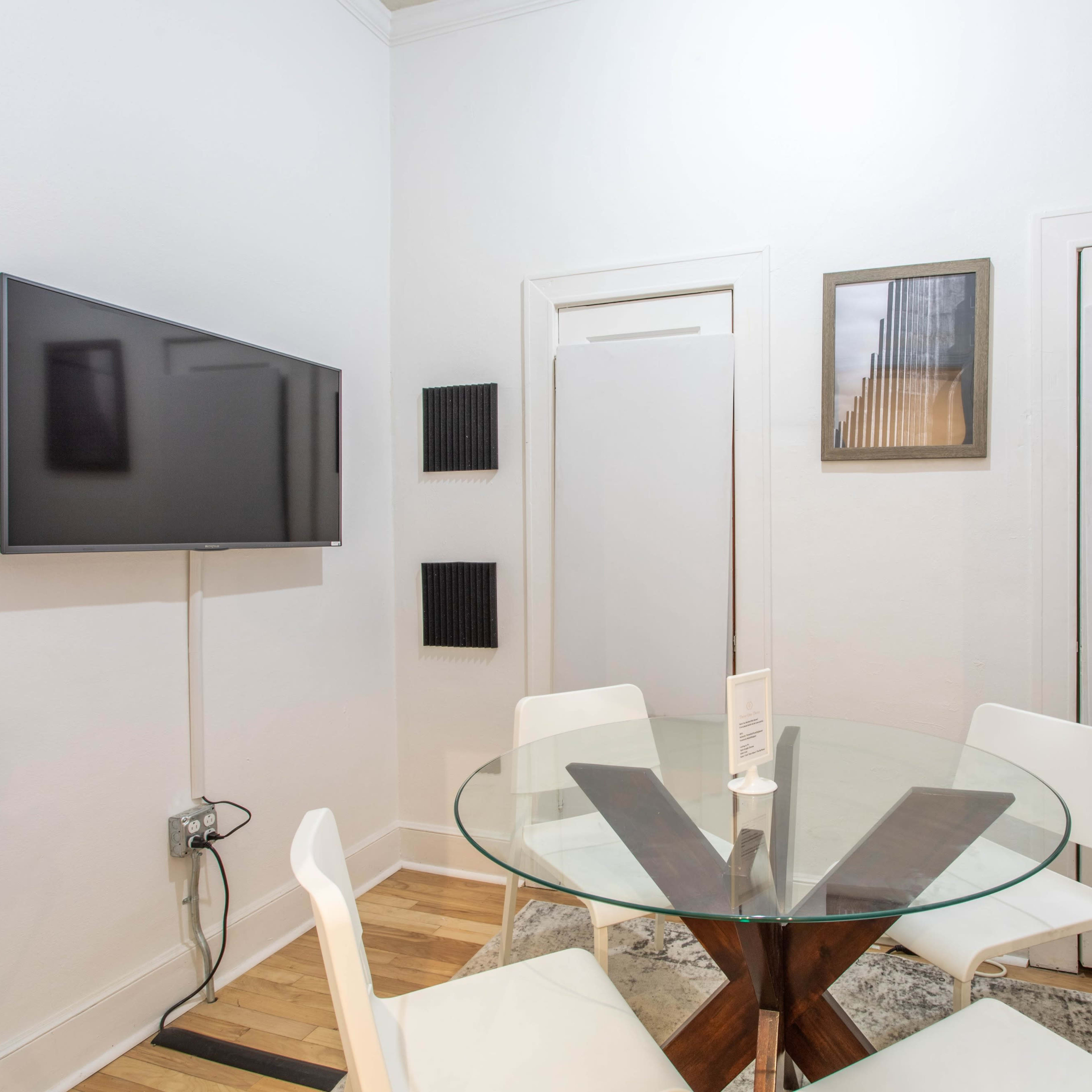 A round glass dining table with white chairs is set in a small room featuring a wall-mounted TV and two closed doors.