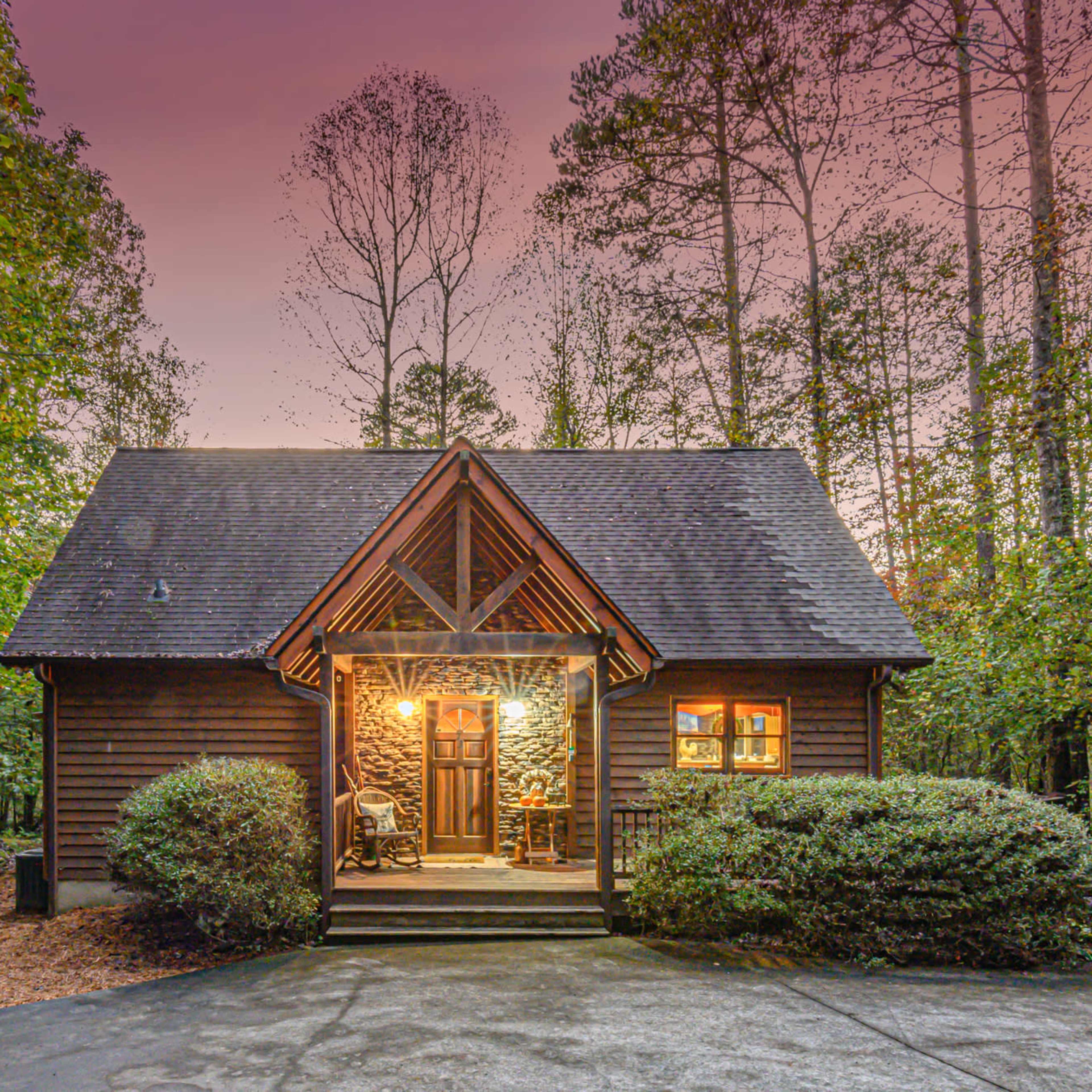 A wooden cabin with a sloped roof is situated amid trees and shrubs, featuring a welcoming front porch and a warmly lit entrance.