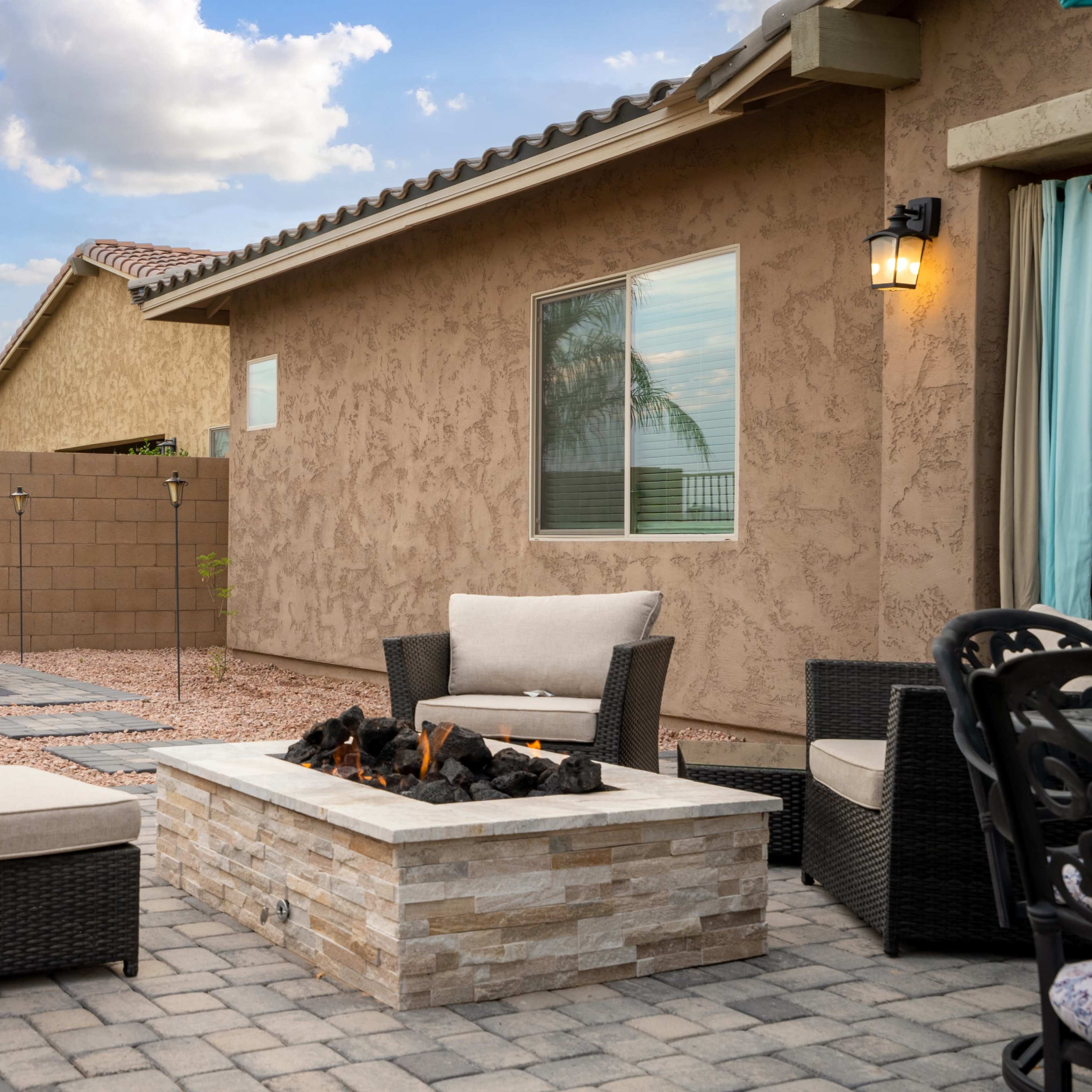A backyard patio features a stone fire pit surrounded by wicker furniture, with a hot tub and a wall of plants in the background.