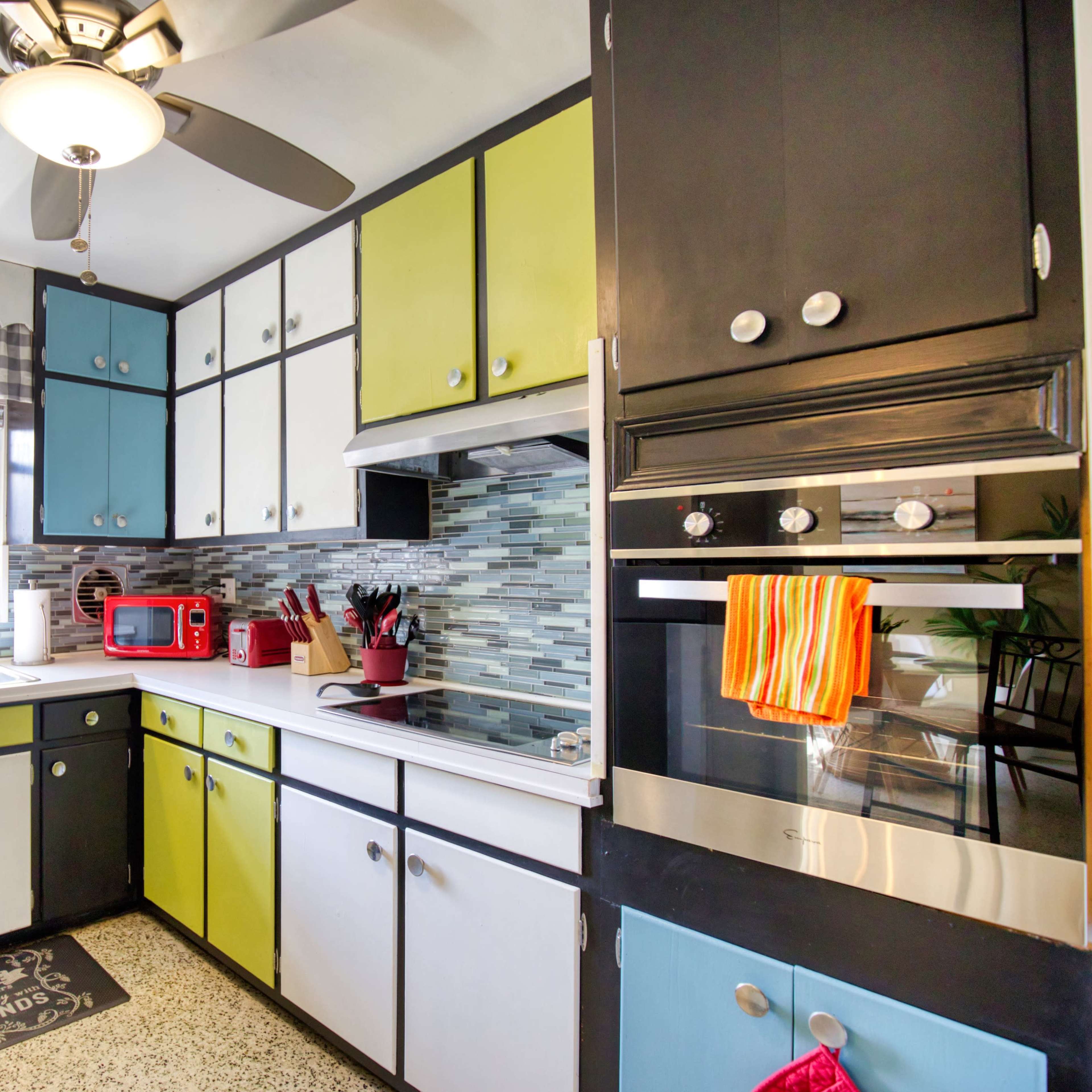The kitchen features a mix of black, white, and brightly colored cabinets, with a window above the sink and various utensils arranged on the countertop.