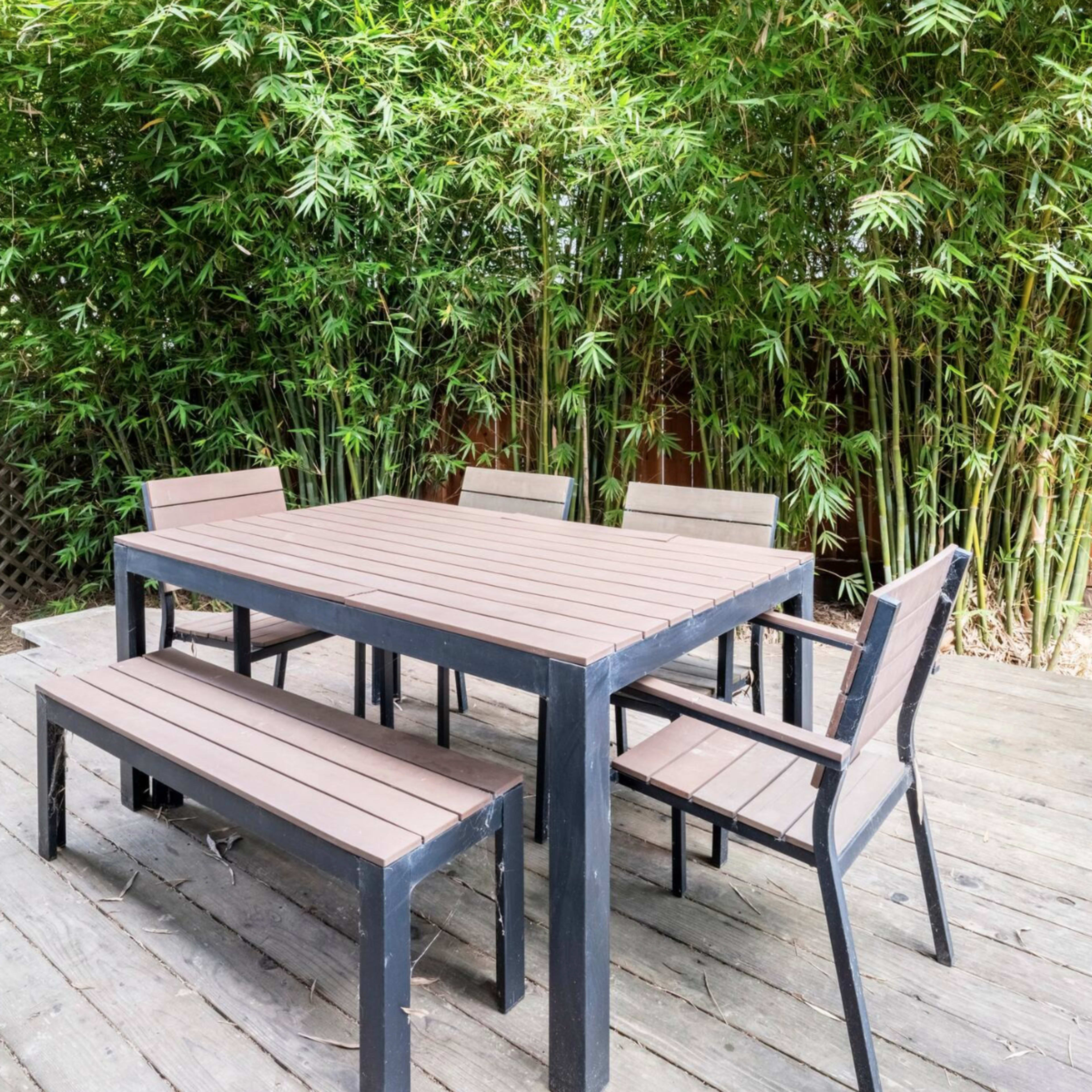 A wooden outdoor dining set is placed on a deck surrounded by tall bamboo plants.