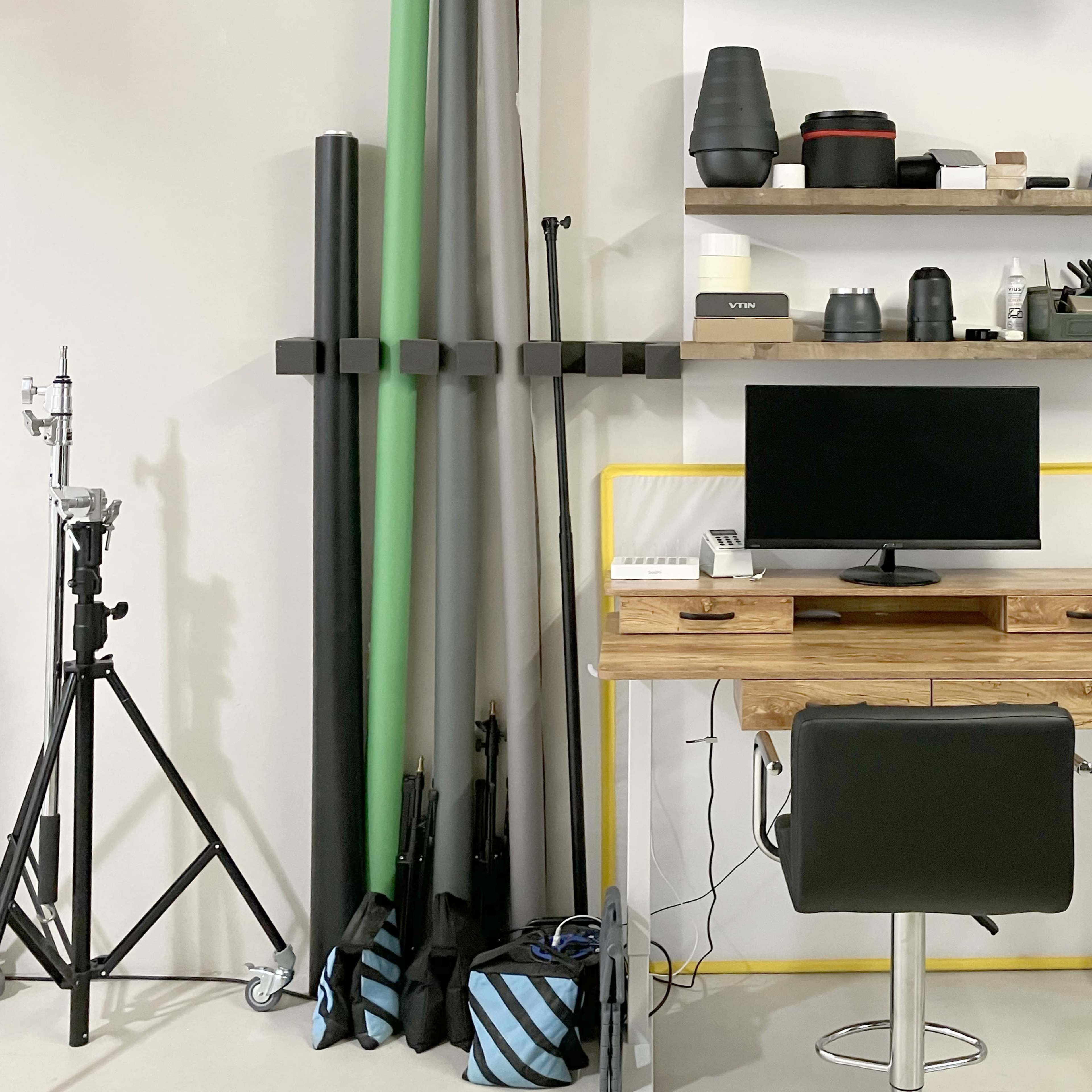 A minimalist workspace with a wooden desk, a monitor on a stand, and various organizational items on shelves, alongside photography equipment arranged against a wall.