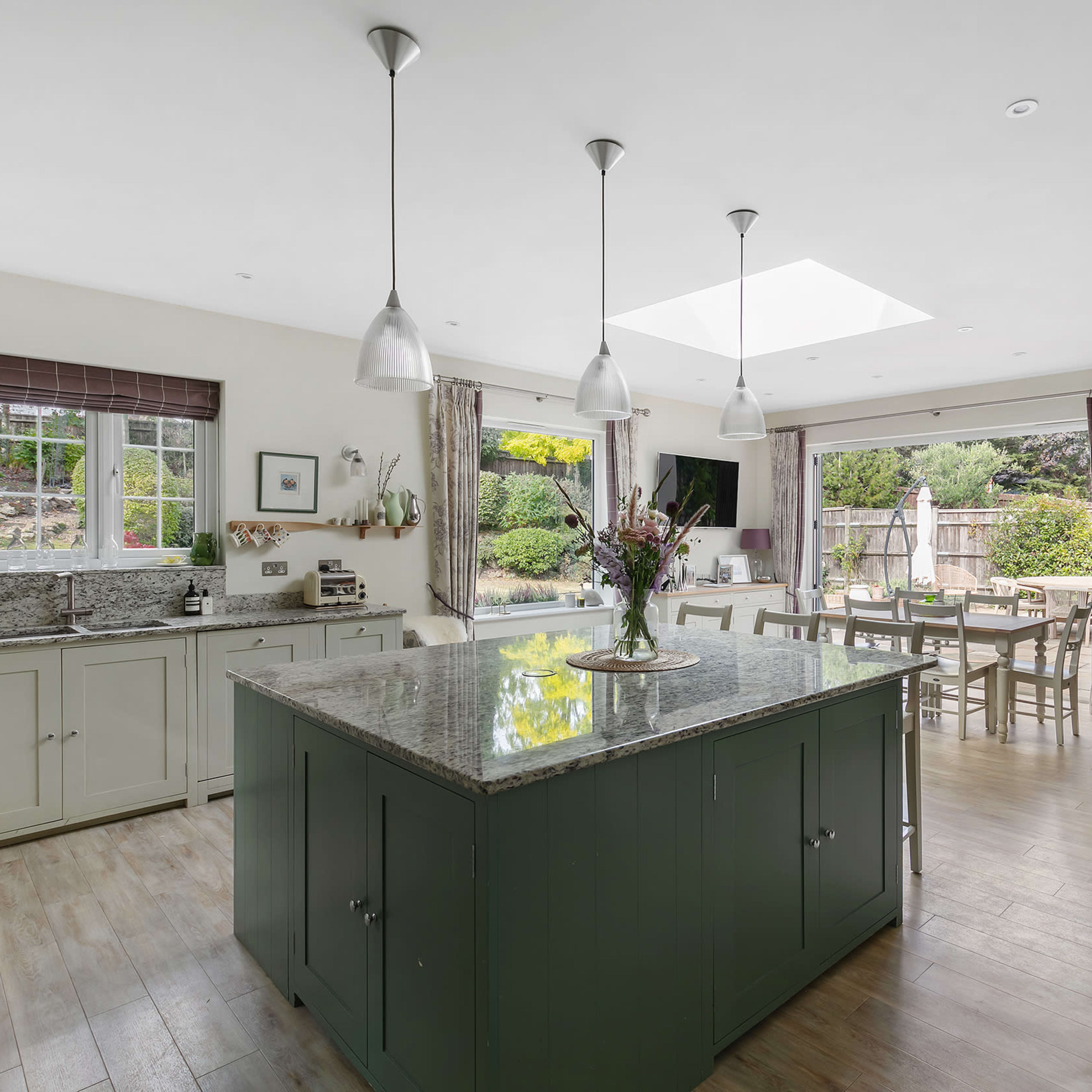 A spacious kitchen with green cabinetry features a large island and opens to a dining area with a table and chairs, illuminated by natural light from a skylight.