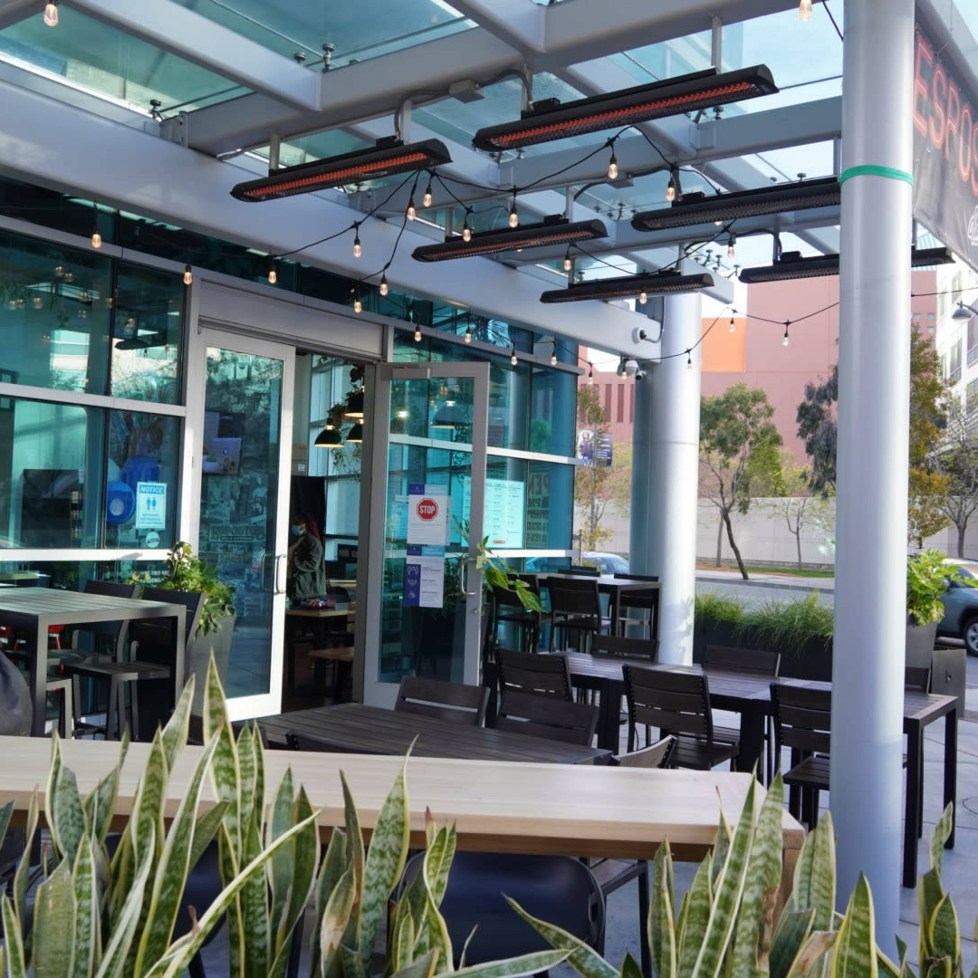 A modern outdoor dining area features tables, chairs, and hanging lights beneath a glass roof with greenery along the edges.