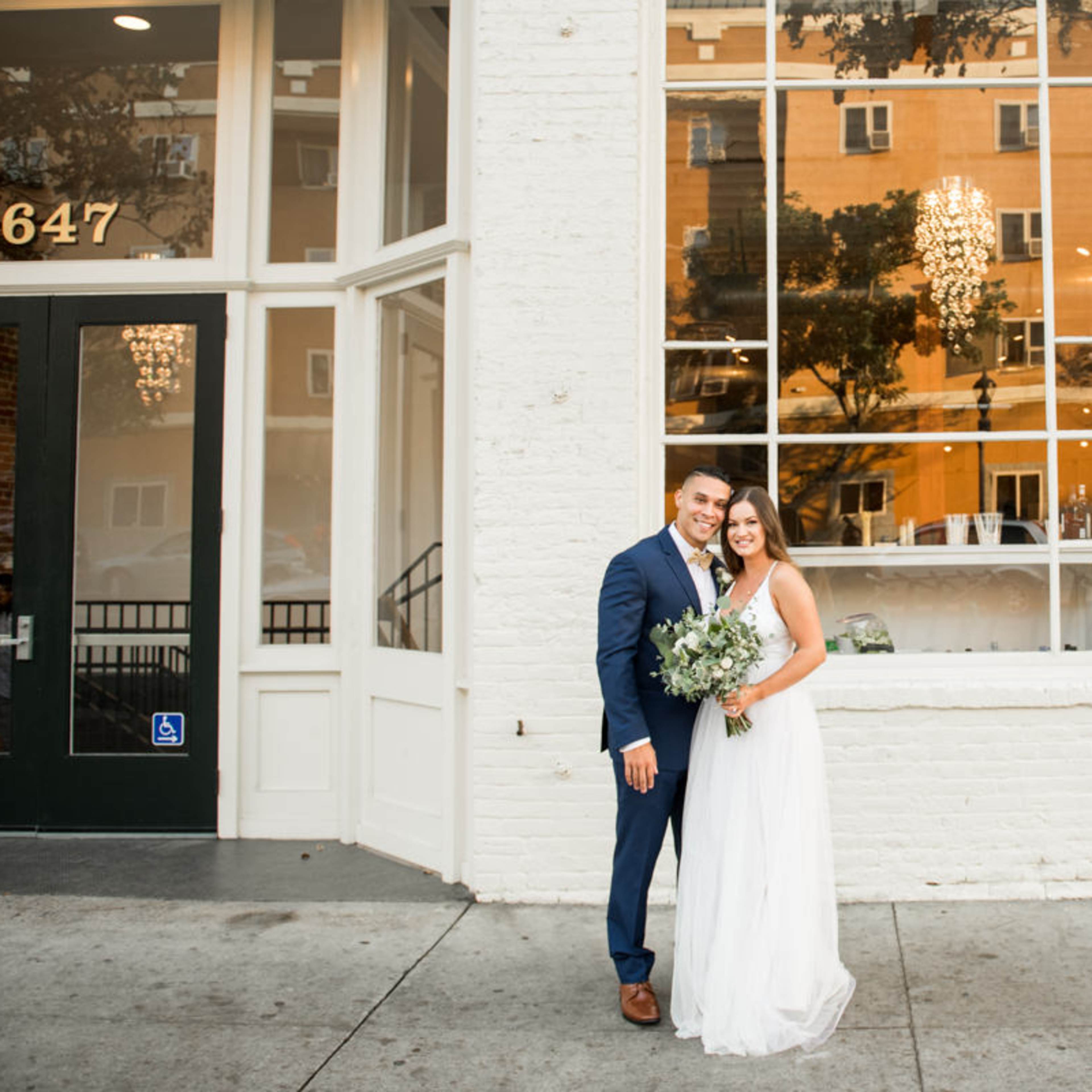 A couple in formal attire stands outside a white brick building with large windows, holding a bouquet of flowers.