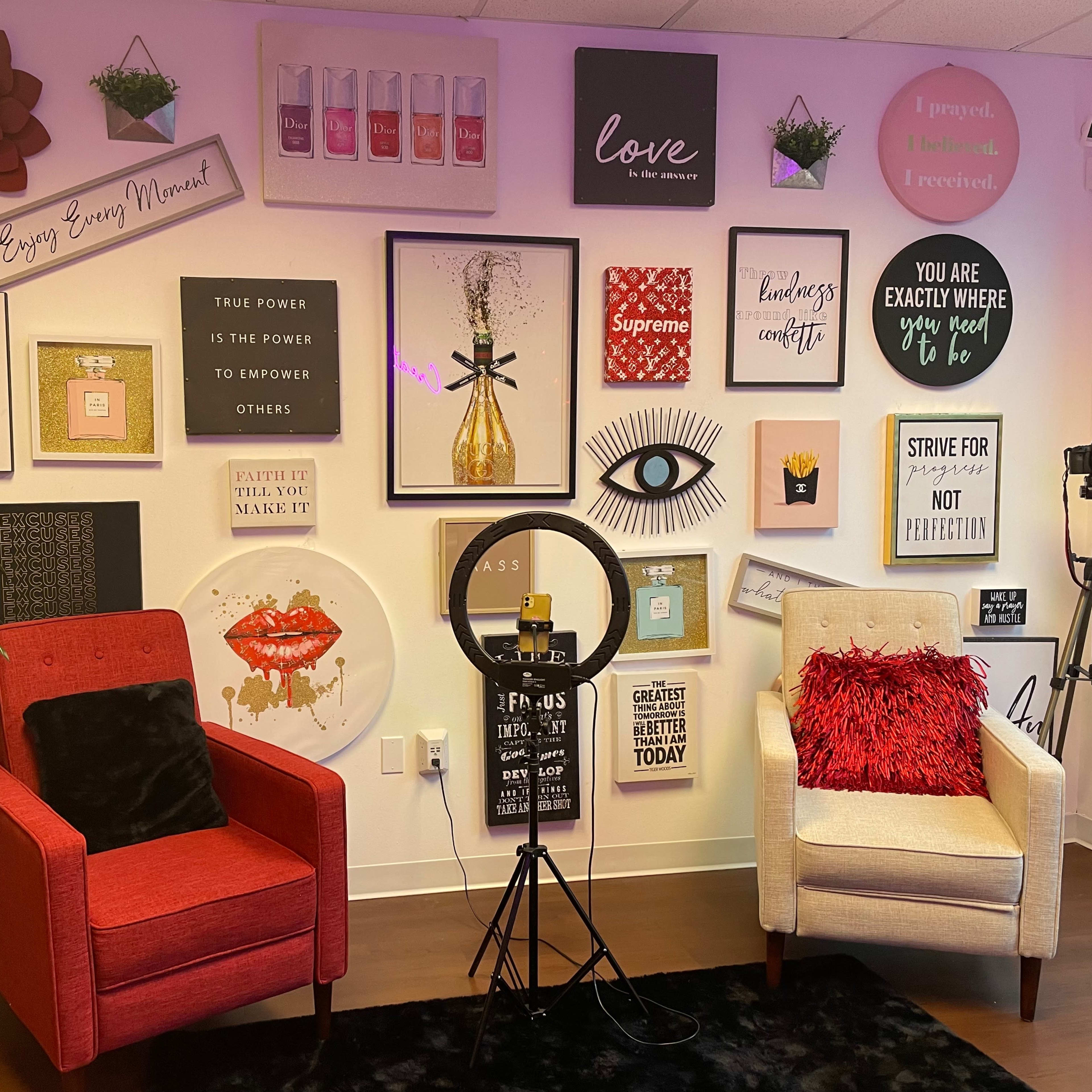 A decorated wall with various motivational posters, two upholstered armchairs, a ring light, and a camera stand.