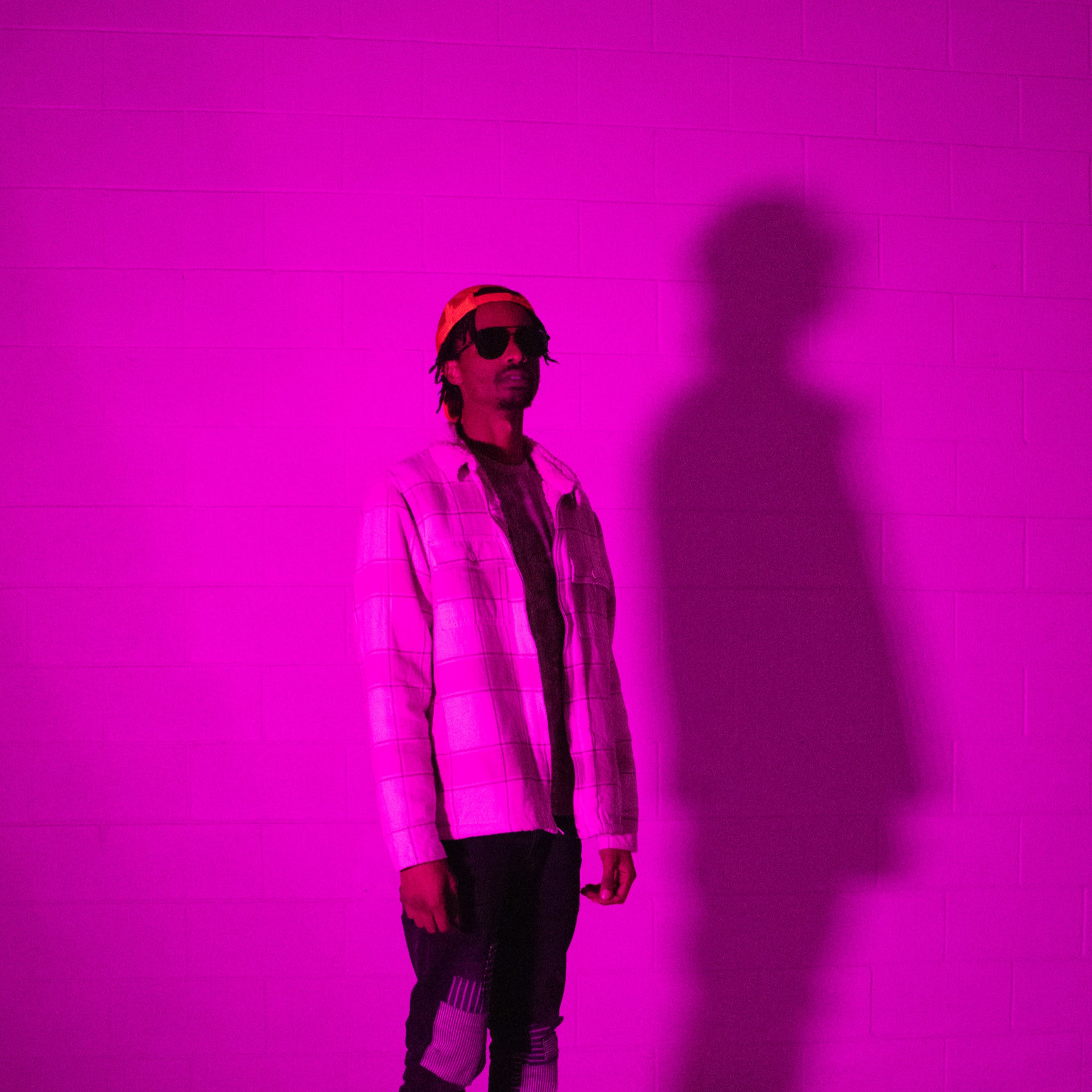 A person wearing sunglasses and a yellow hat stands against a pink wall, casting a shadow behind them.