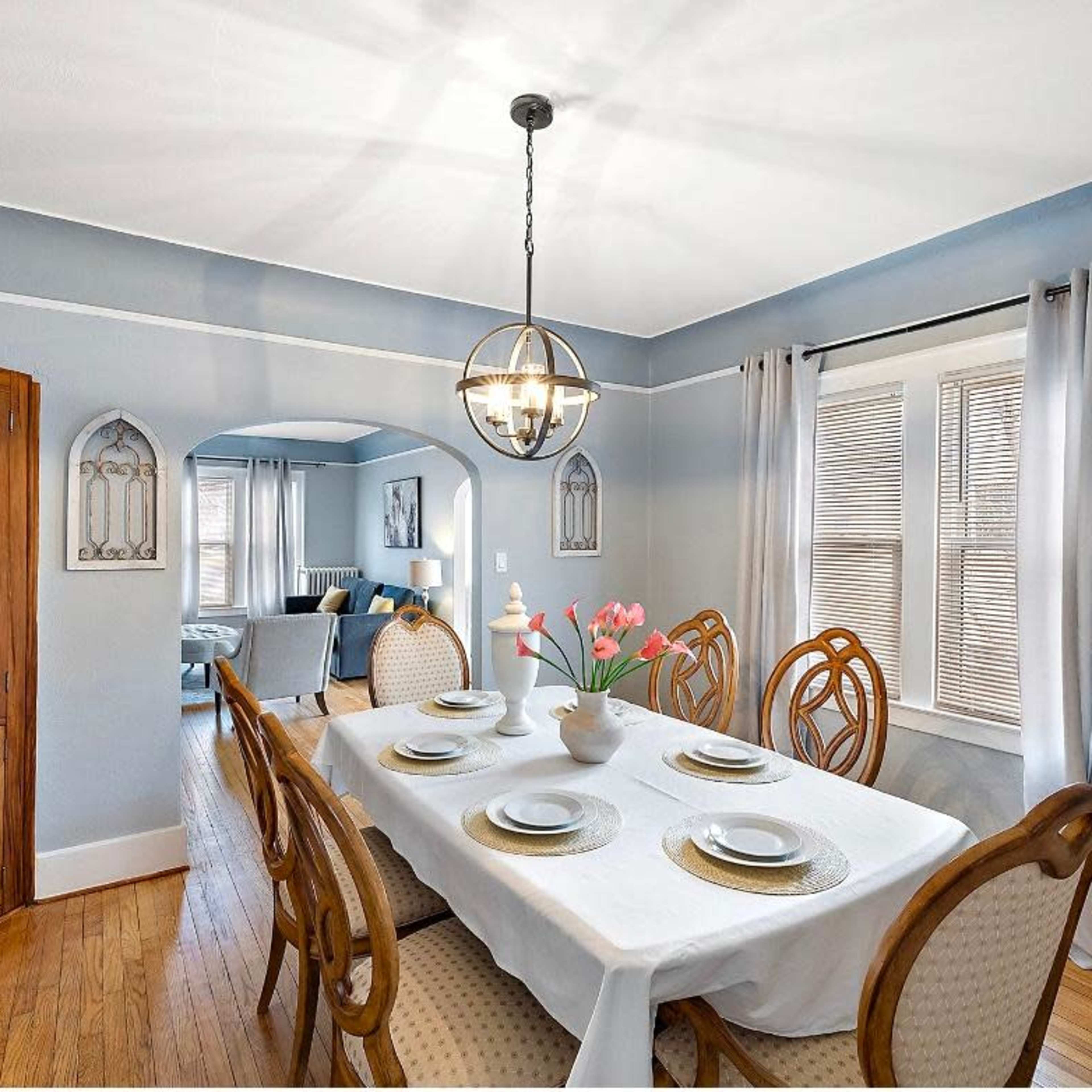 A dining room features a table set for six, surrounded by wooden chairs, with a lit pendant light overhead and a glass cabinet against the wall.